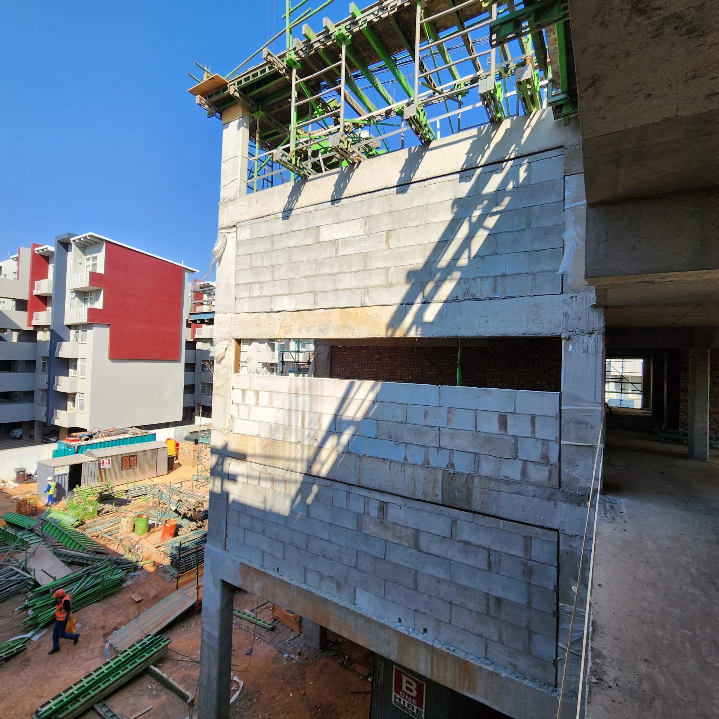 A block infill panel seen from an upper level, courses running from the beam soffit down past a horizontal slot opening, with the site yard and the neighbouring blocks behind.
