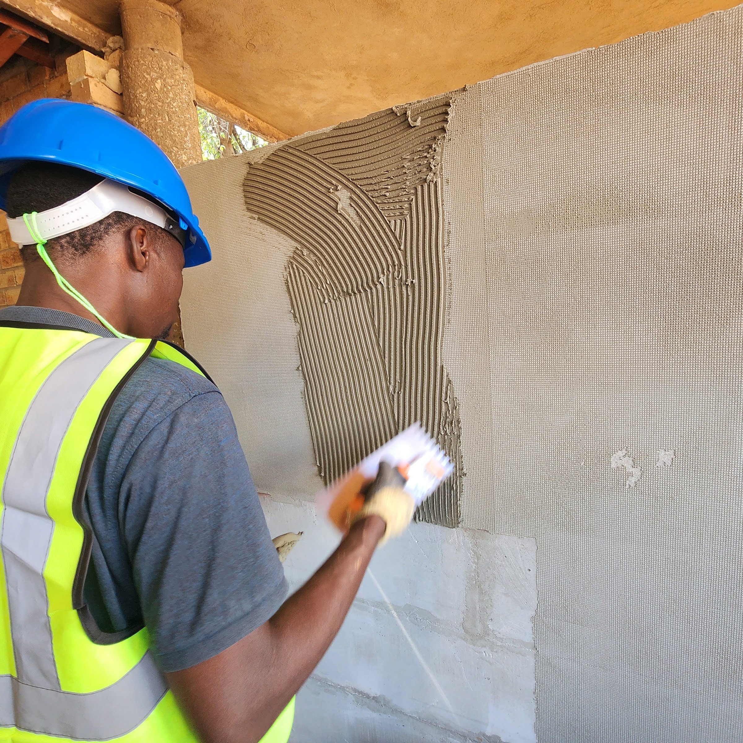 A worker combs adhesive onto the wall with a notched trowel. The serrated ribbons stand up crisply where they run out onto a face on which the square grid of embedded reinforcing mesh still shows through the coat.