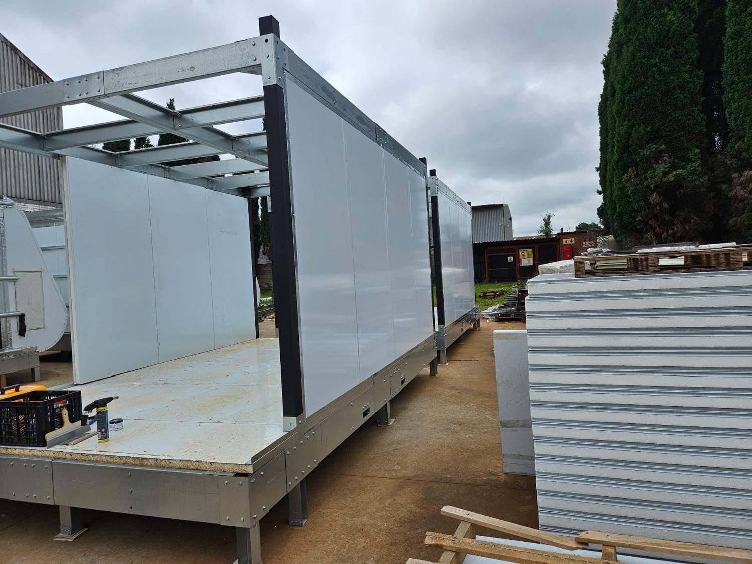 The system, mid-assembly. Cold-formed roof grid and corner posts up; LiteSpan wall boards standing between the posts. The stack of boards on the right shows the cut edge — two steel skins, EPS core — which is the same board already standing as the wall.