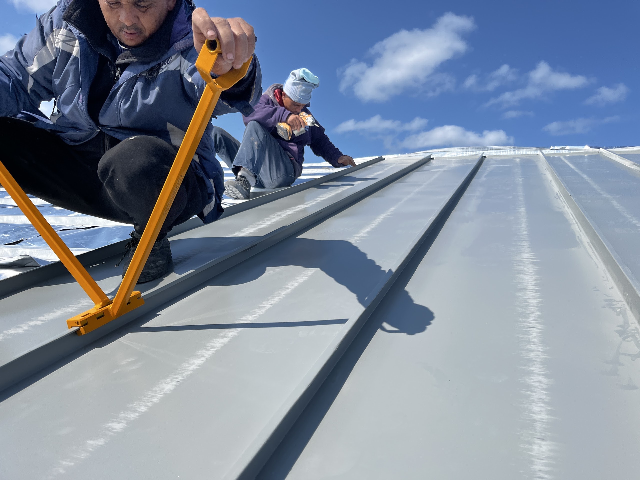 Hand-seaming the roof. One roofer walks a yellow seaming tool along a standing seam while a second fixes the next sheet down; the silver reflective foil laid over the rafters shows in the strip still open beyond them.