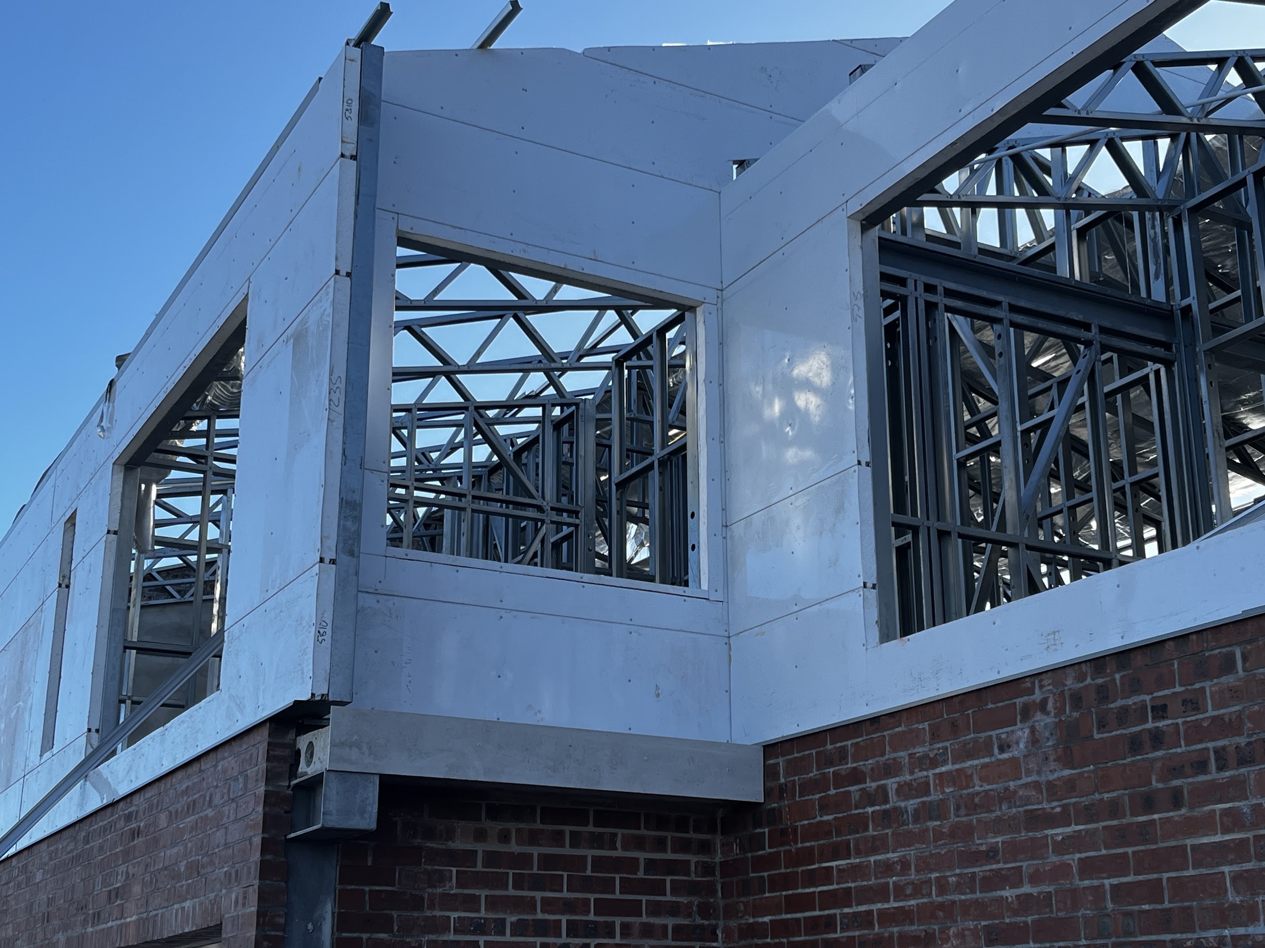 The panel turns the external corner in one plane. Through the two window openings the frame behind is completely visible - studs, K-bracing, lattice trusses and reflective foil - and none of it interrupts the outer face.