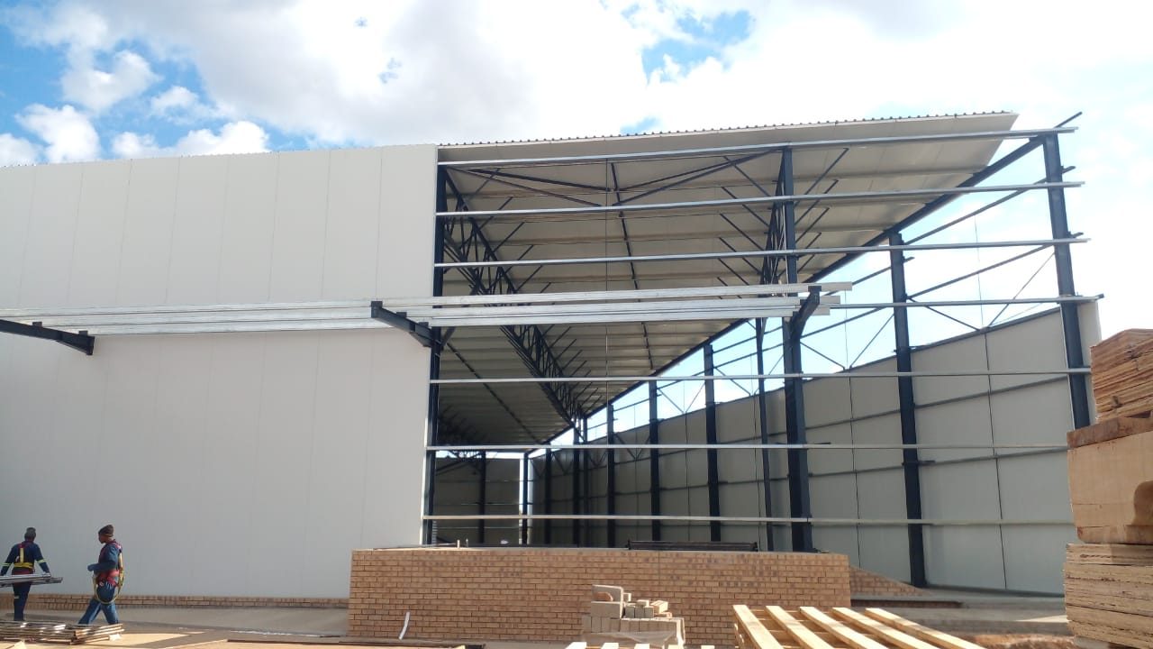 Installation sequence, in one photograph. The wall panel is going straight onto the bare steel frame — two men carrying a board between them at the left, nothing behind it. Frame, panel, done: cladding, insulation and internal finish are a single fix.