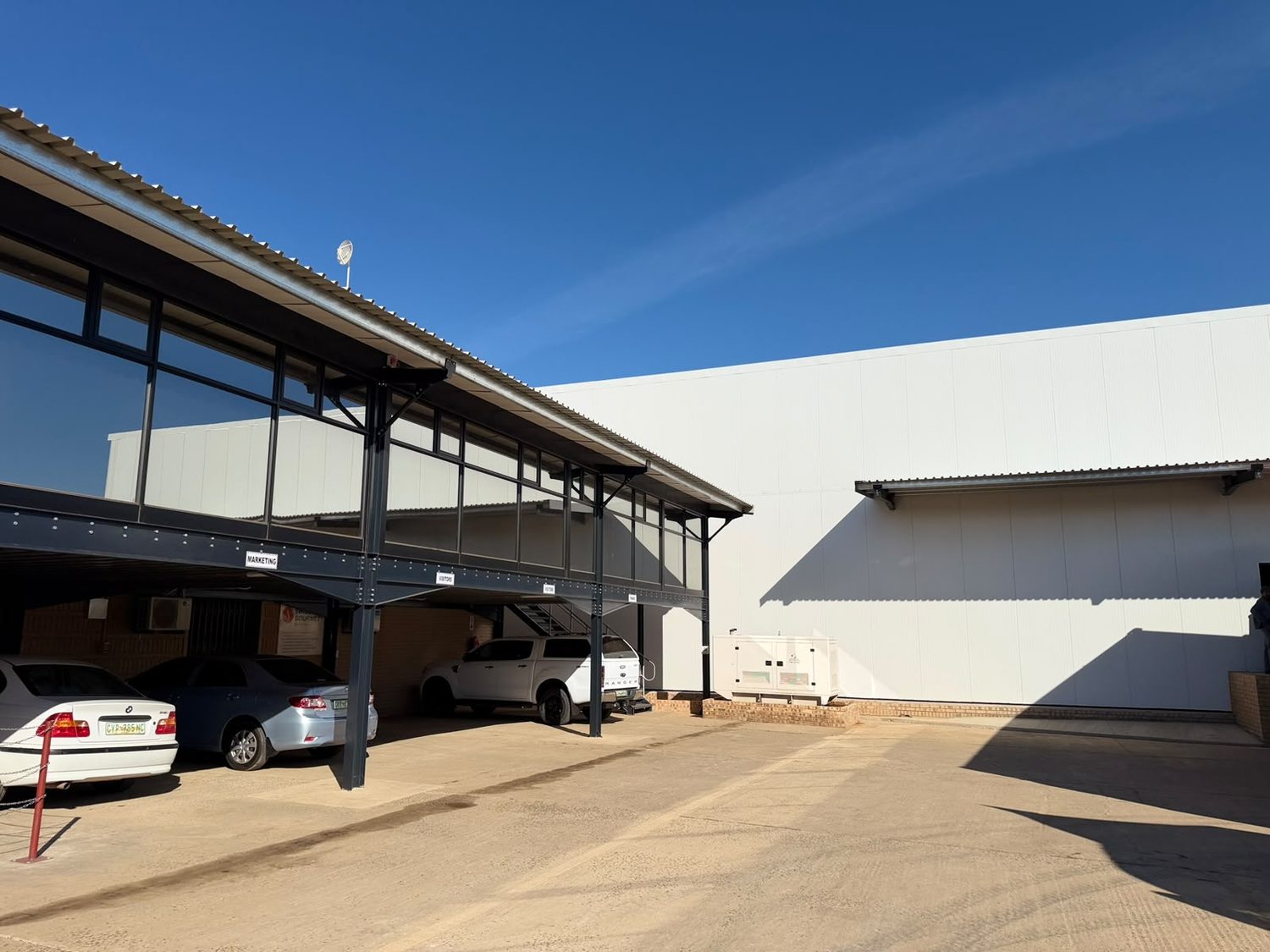 Where the store meets the office block: the panel wall lands on a brick plinth, and the canopy roof, gutter and downpipe are flashed into the panel face. A flat, joint-lined wall that takes the rainwater goods directly and needs no paint system.
