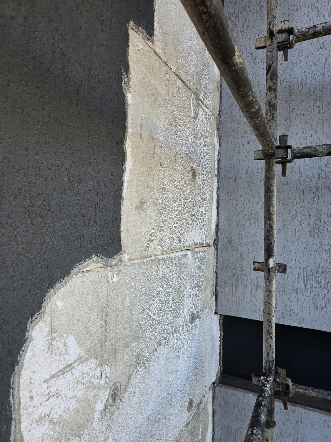 Three layers of the system in one frame: finished charcoal textured topcoat on the left, the grey basecoat being floated out in the middle, and the bare dowelled EPS board still showing. Scaffold standard running past it — a real wall, not a sample panel.