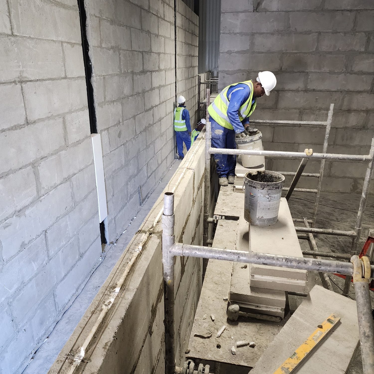 Laying. Blocks are buttered and set in thin-bed adhesive from a bucket, and the ring-beam reinforcement runs along the top of the wall - the wall is built up to and tied into the frame, which carries everything.