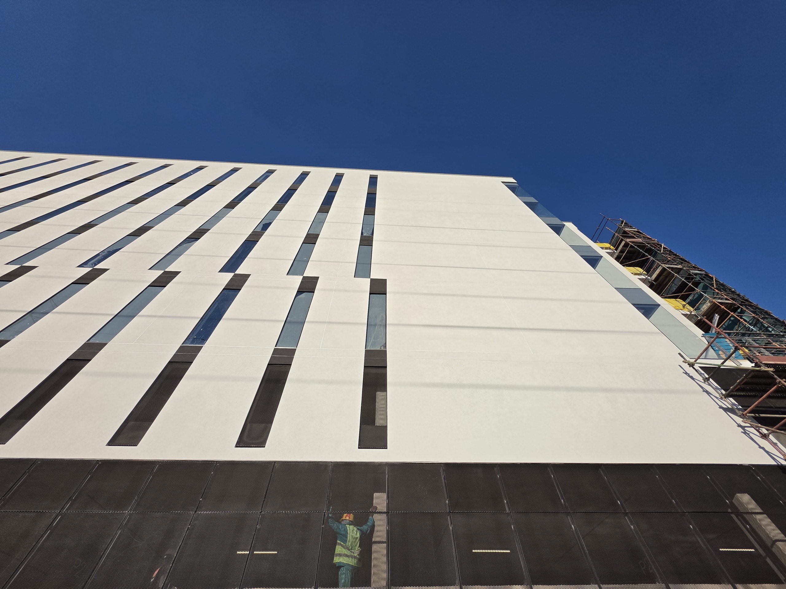 Looking along the finished elevation in raking sun. The flat white planes read as one surface between the slot openings, scaffold still up at the far end, a worker in hi-vis at the base for scale.