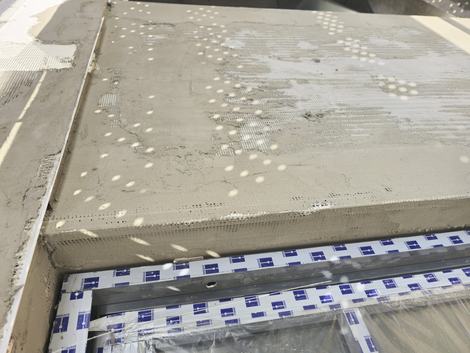 Terramesh embedded in the wet Styrobond basecoat, carried over the window head and returned down into the reveal. The mesh is visible through the basecoat exactly where facades usually crack — at the opening corners and the arris — because the reinforcement is wrapped, not butted.