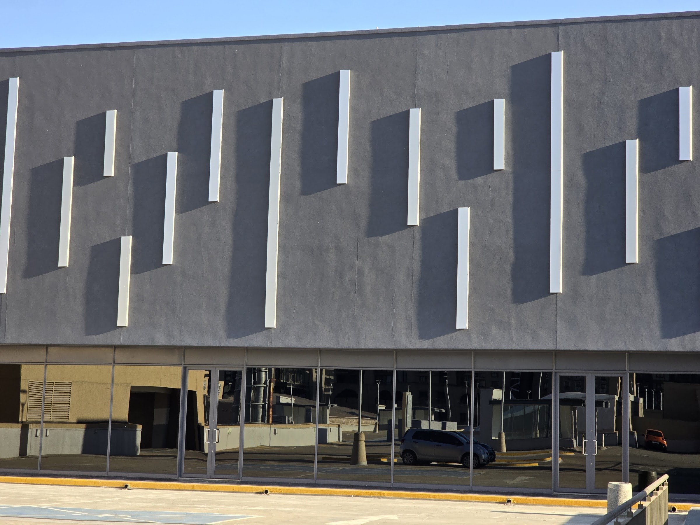 Low sun rakes across the rendered facade and every white vertical strip throws a hard shadow onto the textured face; glazed shopfront and parking deck below.