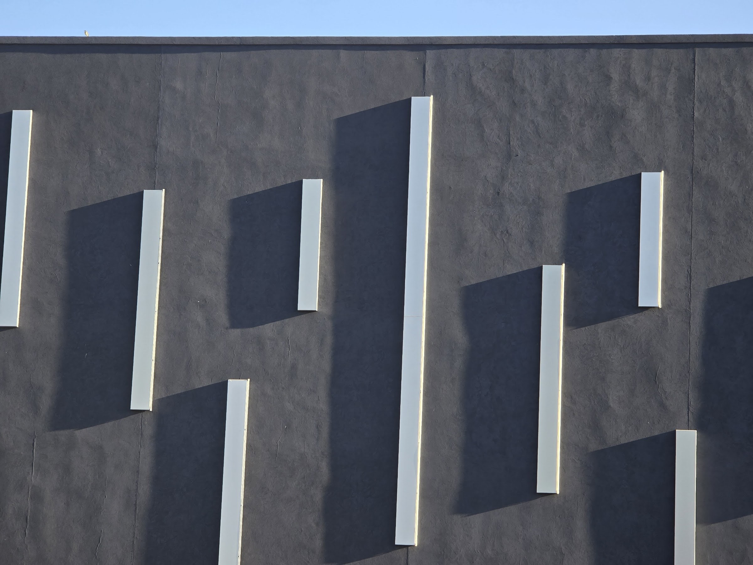 Close view of the completed dark render with the white profiled strips set off the surface, each casting a shadow across the textured face, with a strip of sky above the parapet.