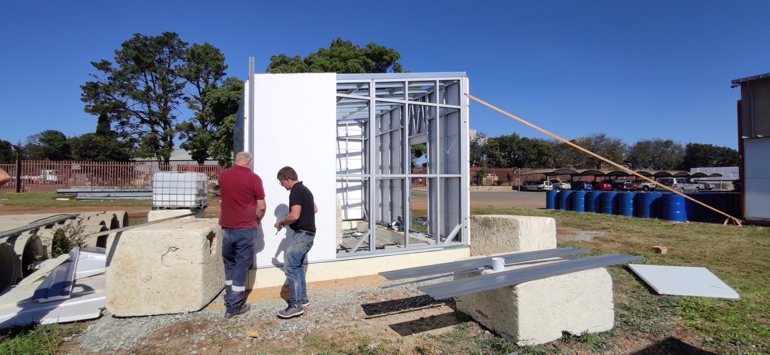 Half frame, half envelope. Knee-braced cold-formed steel studs with a trimmed opening on the right; EPS board already closing the wall on the left. Two people, no crane, no wet trade on the wall.