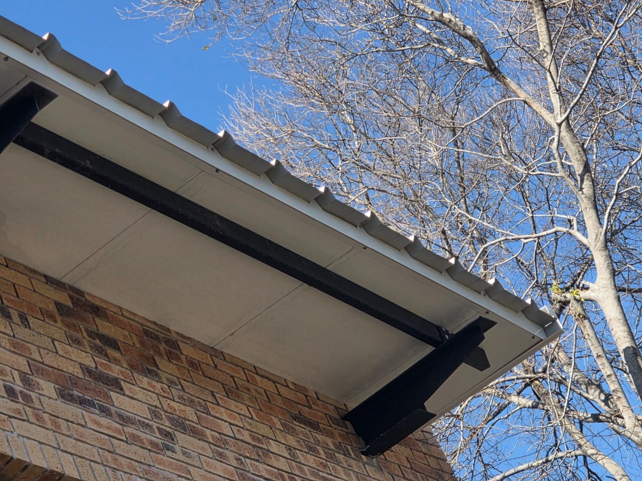 Under the eave at a corner. The flat white pan is the finished surface you see from the ground; the ribs of the top skin run along the open edge above it, and a black steel cantilever bracket carries the overhang out past the brickwork. The straight lines across the pan are the joints between panels.