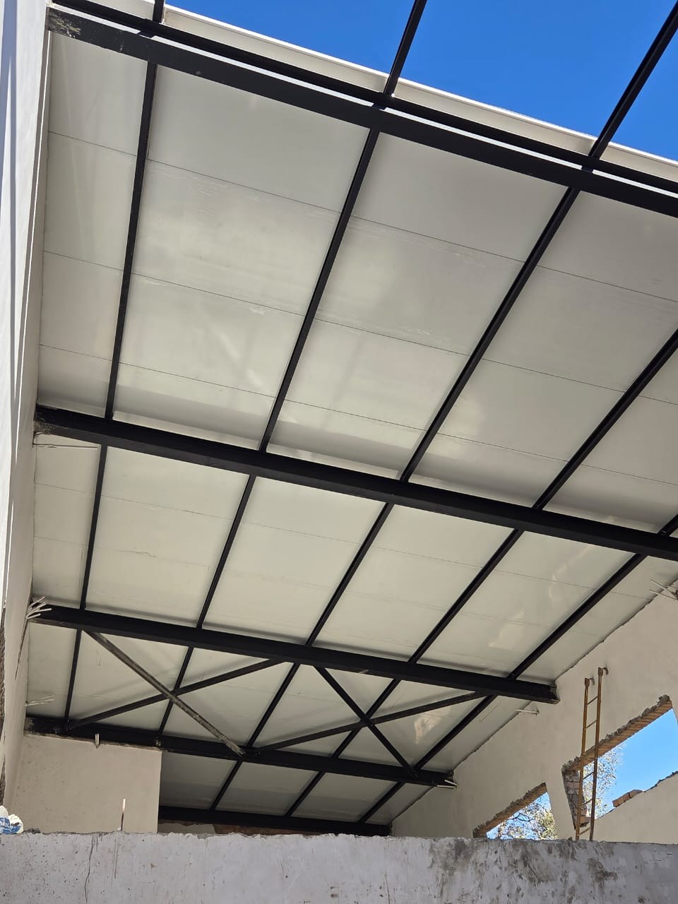 The same panel from inside, looking up. The white pan spans between the exposed black purlins and is the finished ceiling - nothing is fixed below it. Rafter cross-bracing crosses it at the lower end, and one bay at the top is still open to the sky.