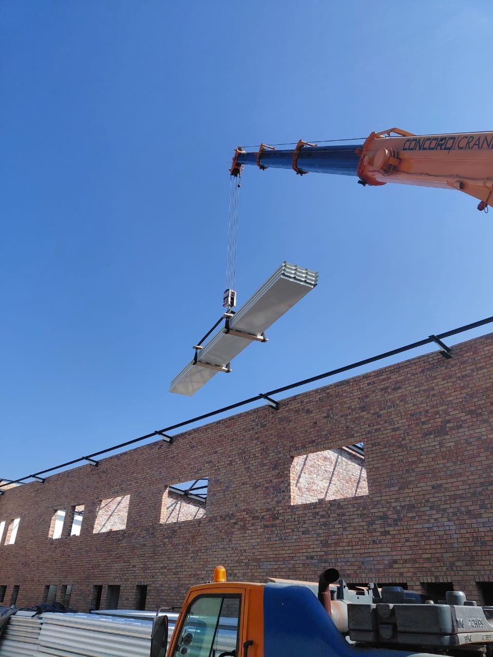A bundle of nested panels slung on a spreader beam under a truck crane, swung in over the brick wall with the black steel purlins already fixed along it.