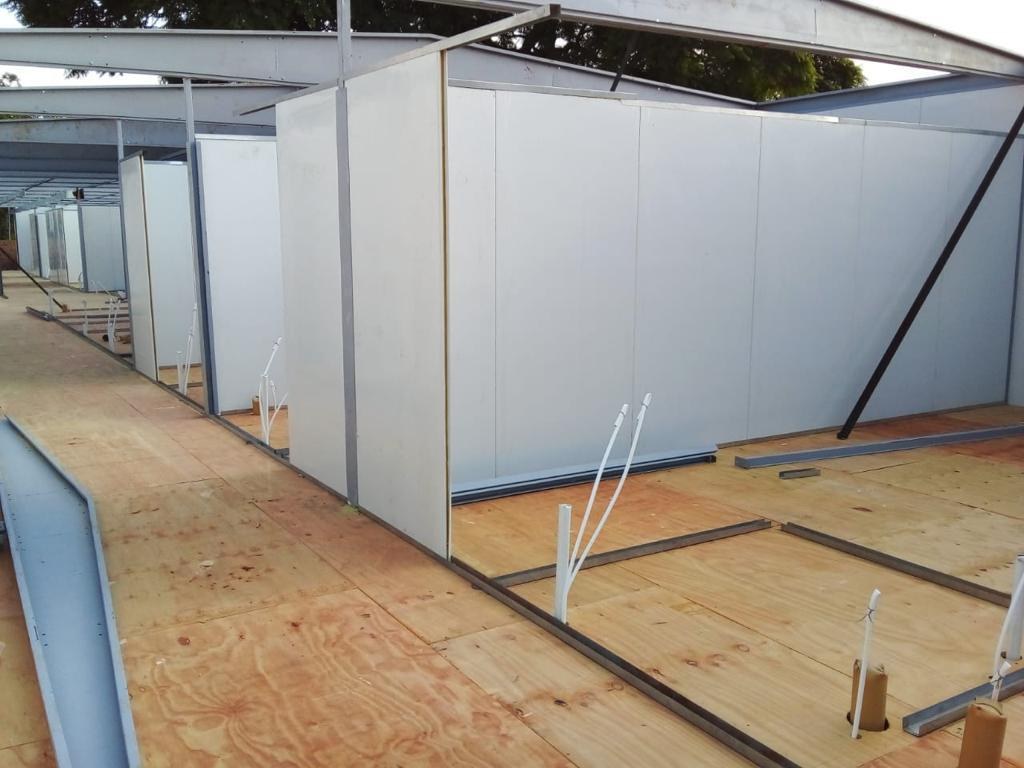 Down the length of the deck: panel walls forming a run of rooms. At the external corner the cut edge of the panel reads as a thin tan line, the base sits in a steel floor track laid on the plywood, and white plumbing stubs stand on wall lines that have no wall yet.