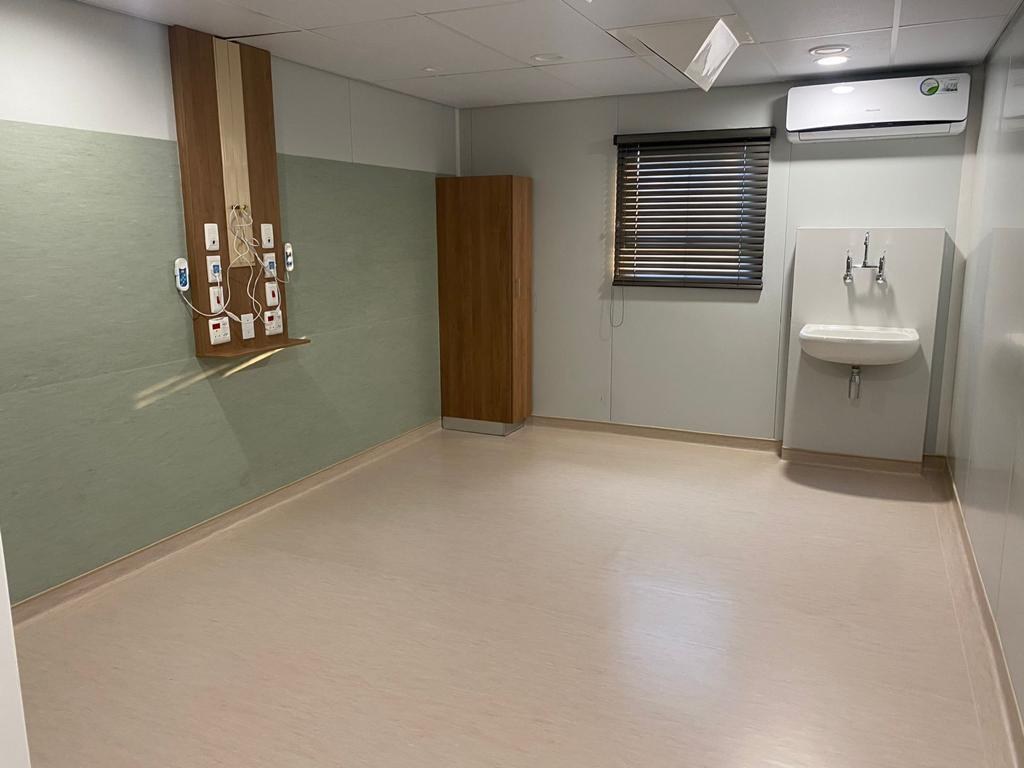 A finished room with the services carried on the panel wall - a timber bedhead board screwed to the panel with service outlets, switch plates and two handsets, a wall basin on a projecting panel opposite, a split air-conditioner, and a blind in the window opening. A green finish runs to about bed height.