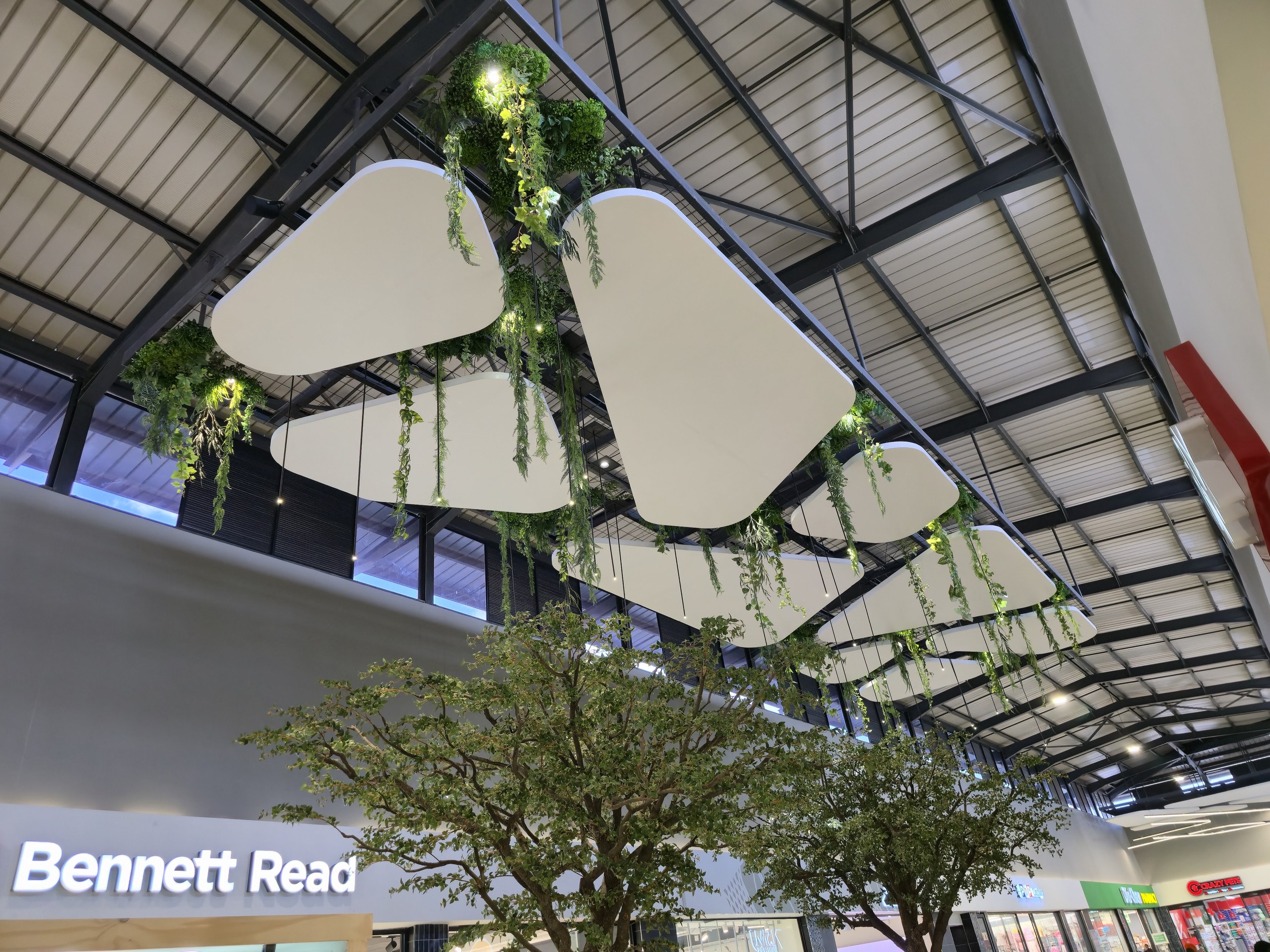 A run of shapes stepping away down the walkway under the exposed steel roof, with garlands of trailing foliage and small warm lights worked in between them and a tree standing in the concourse below.