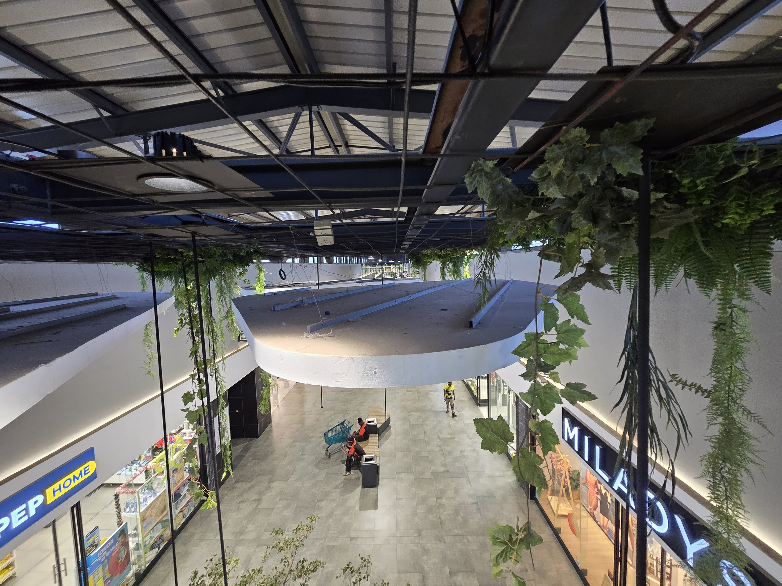 Seen from the upper level, the top of a suspended shape: the top face is left as raw uncoated EPS with light-gauge steel sections laid across it, the coated white edge band wraps the perimeter, and thin black hanger rods run up to the roof structure. Walkway and shopfronts below.