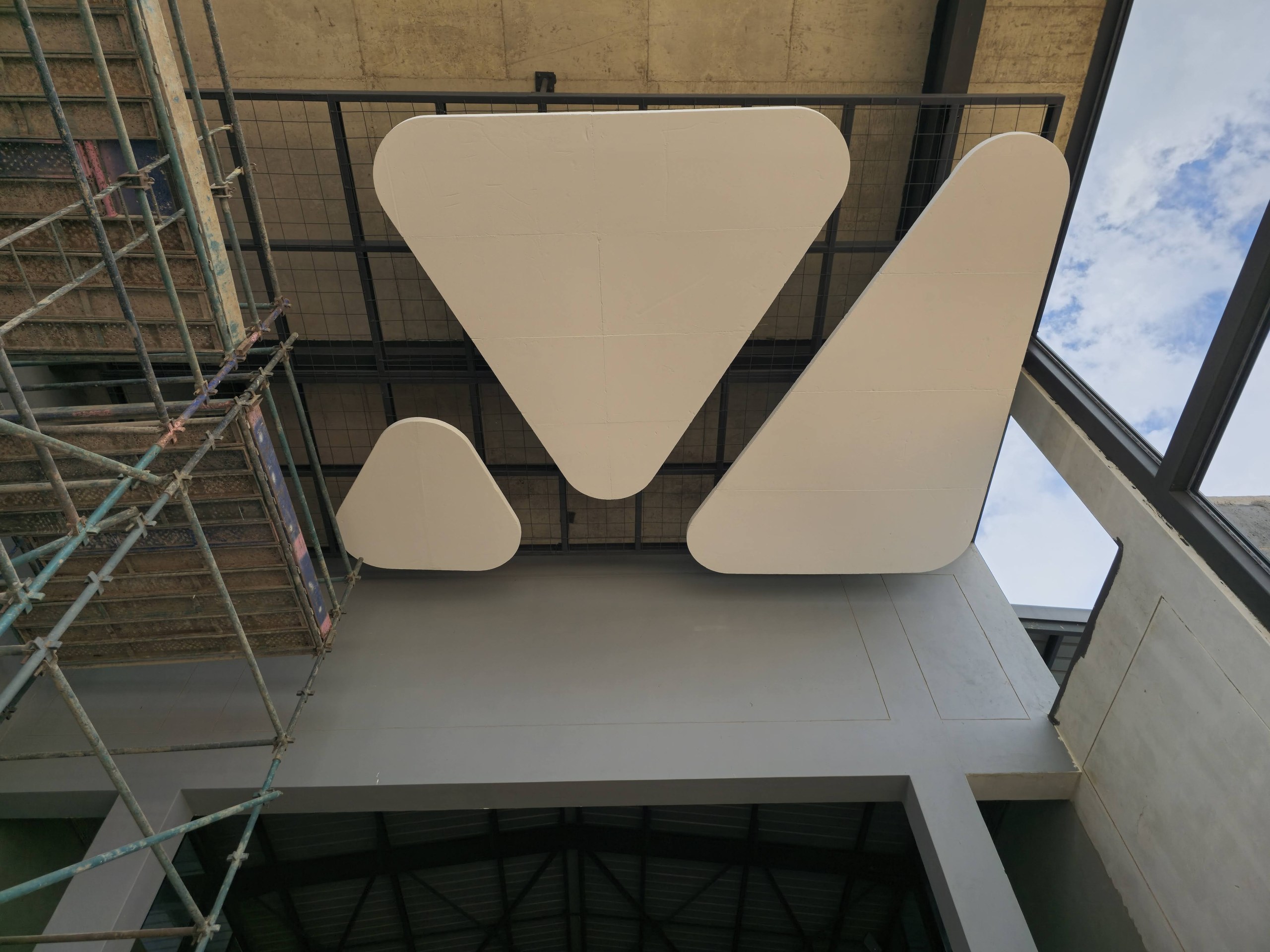 Three coated shapes fixed flat against a soffit above a black steel screen, with scaffold still standing to the left and sky through the glazing to the right - the shapes finished and in place while the surrounding work is still running.