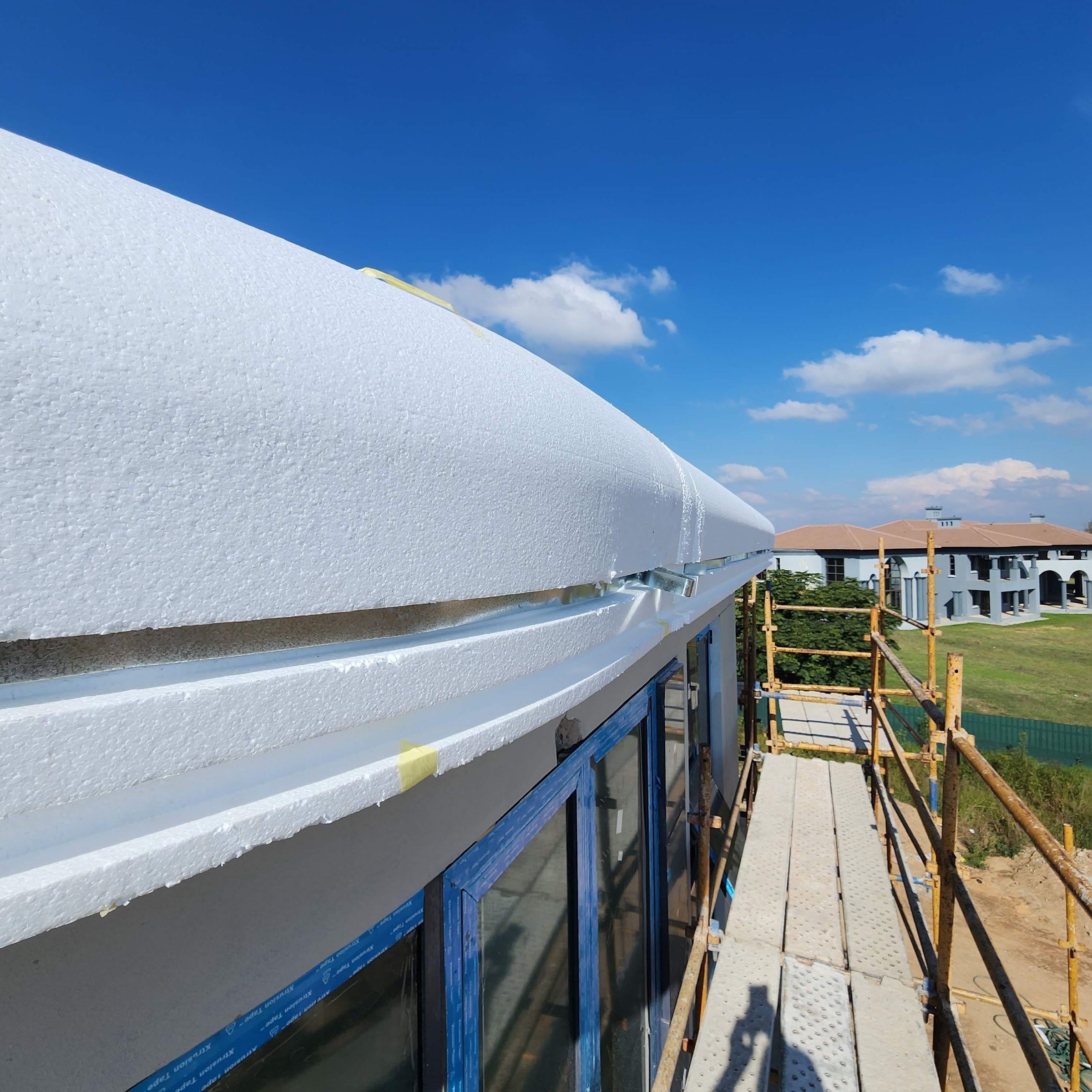 The moulding band in bare expanded polystyrene, fixed above the glazing and running away along the curve of the facade before it is coated - the bead texture is legible on the exposed faces