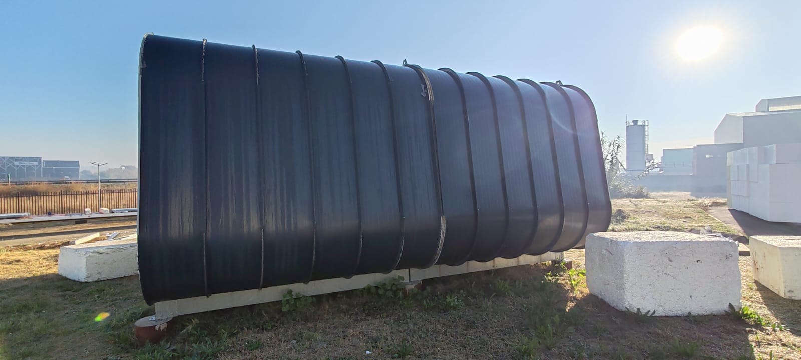 The dark curved shell standing on bearers in the yard, sheet metal wrapped over the barrel form; regularly spaced ridges run over each curve where the sheet is folded.