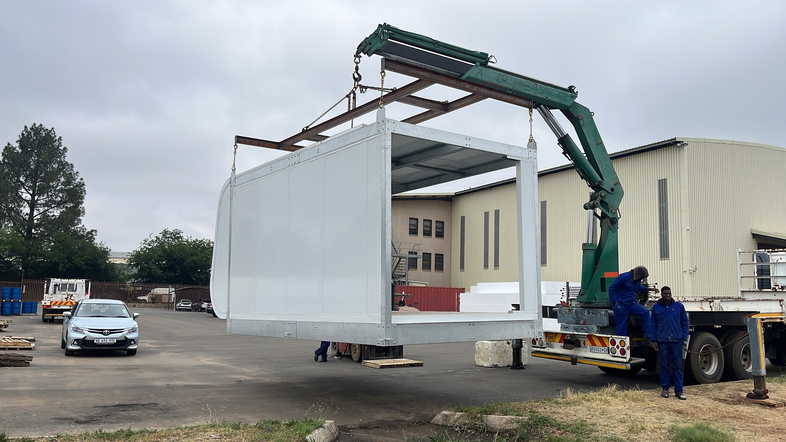 The module slung from a truck-mounted crane on a spreader beam - galvanised perimeter frame, white-faced panels making up the long wall, the rounded end and the open floor deck all legible at once.
