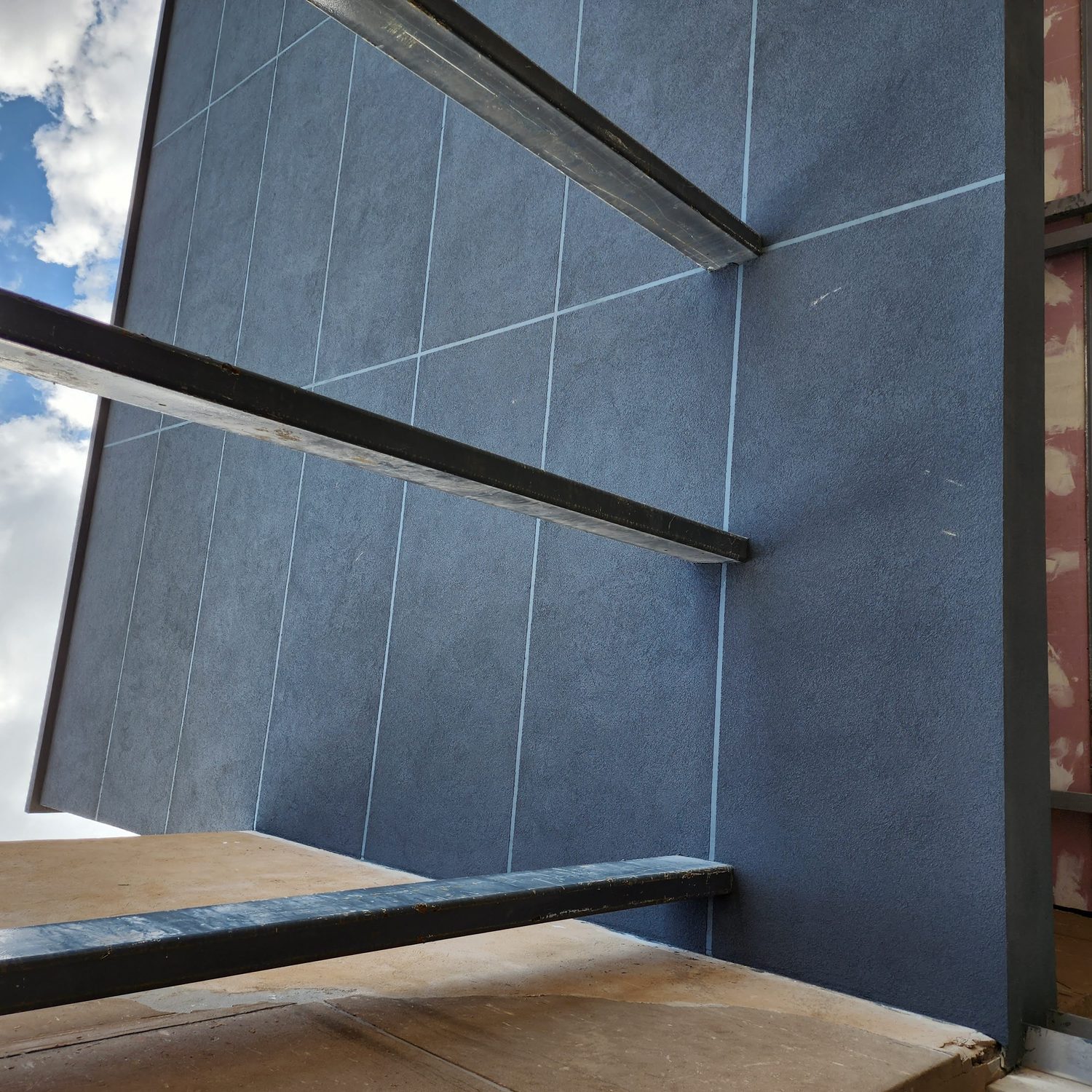 The same coating turns the soffit of the projecting box and runs past the steel columns. Wall and soffit are one skin, so there is no board edge, no trim and no joint to seal where the two meet.