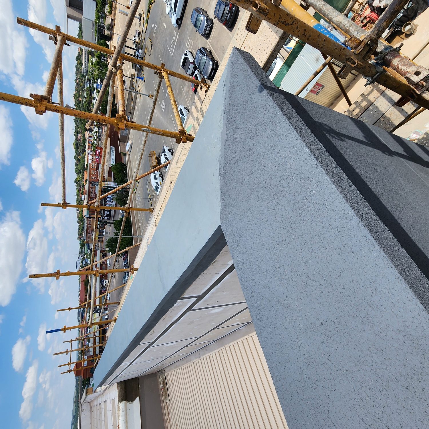 The parapet cap. The render is carried over the top of the parapet and down the far face — no metal coping, therefore no coping fixings penetrating the weather skin at the most exposed line on the building.