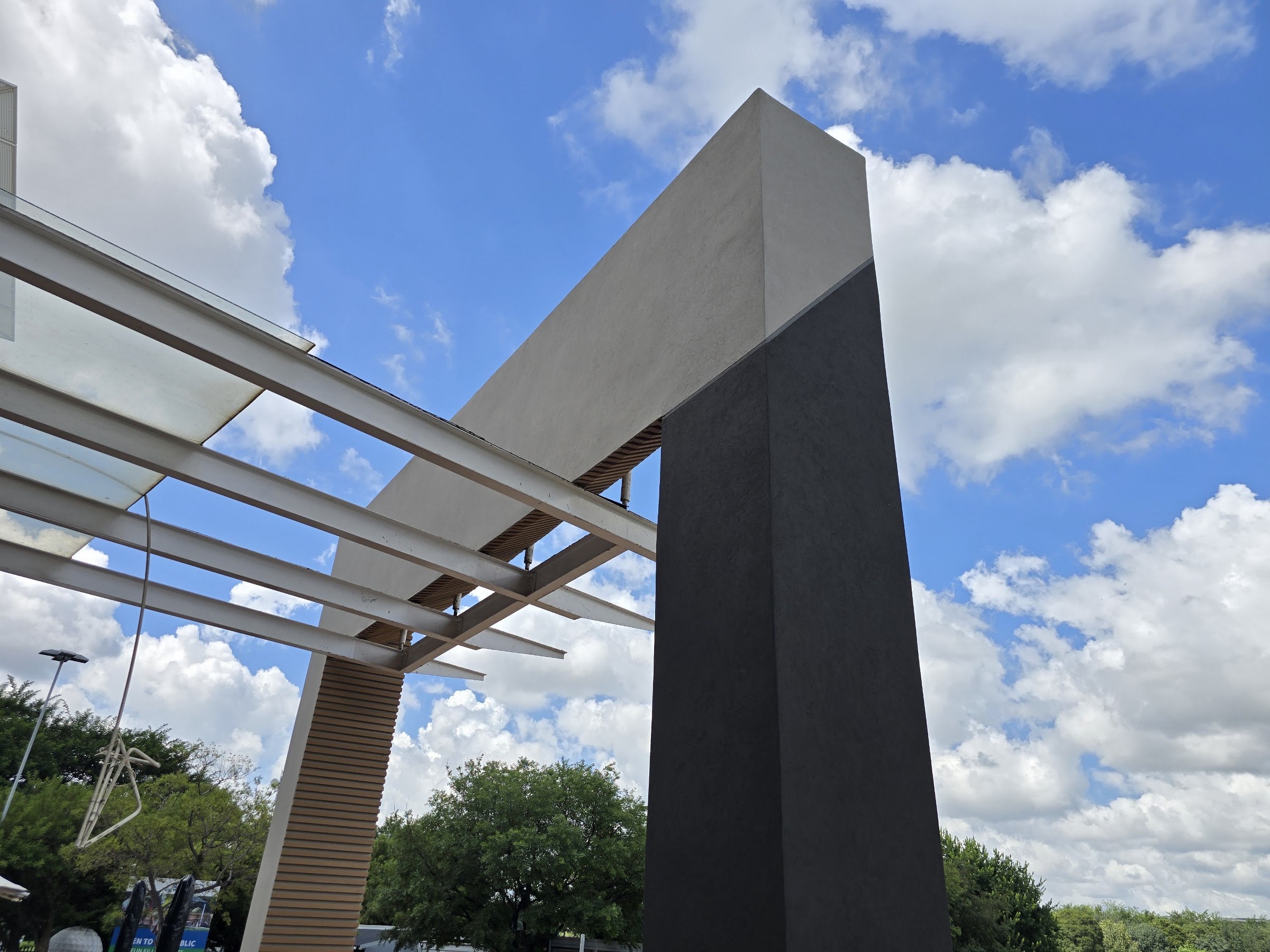 Finished rendered blade against cumulus: a charcoal face with a pale grey top facet, the colour change falling on the facet line rather than on a joint, white steel canopy beams crossing behind, a batten-faced column beyond.