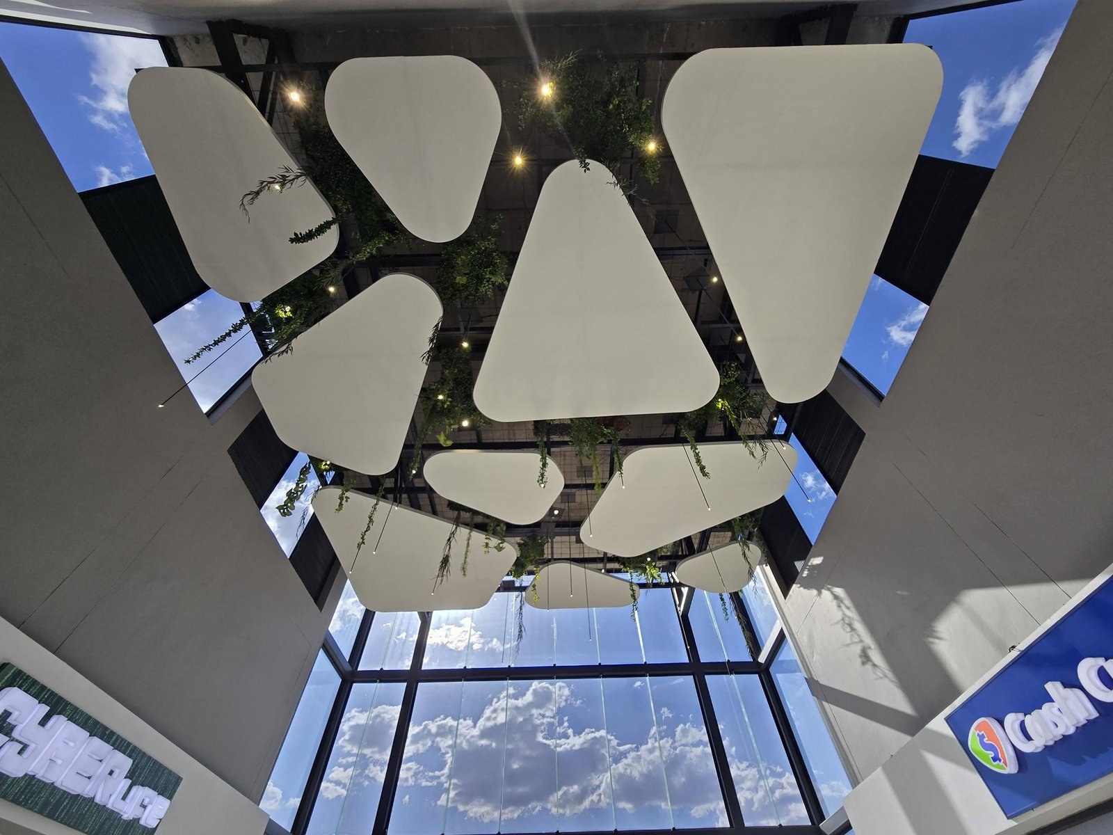 Looking straight up at a mall atrium: flat white shapes with rounded-corner profiles hang on cables below the glazed roof and steel structure, with foliage and downlights threaded between them.