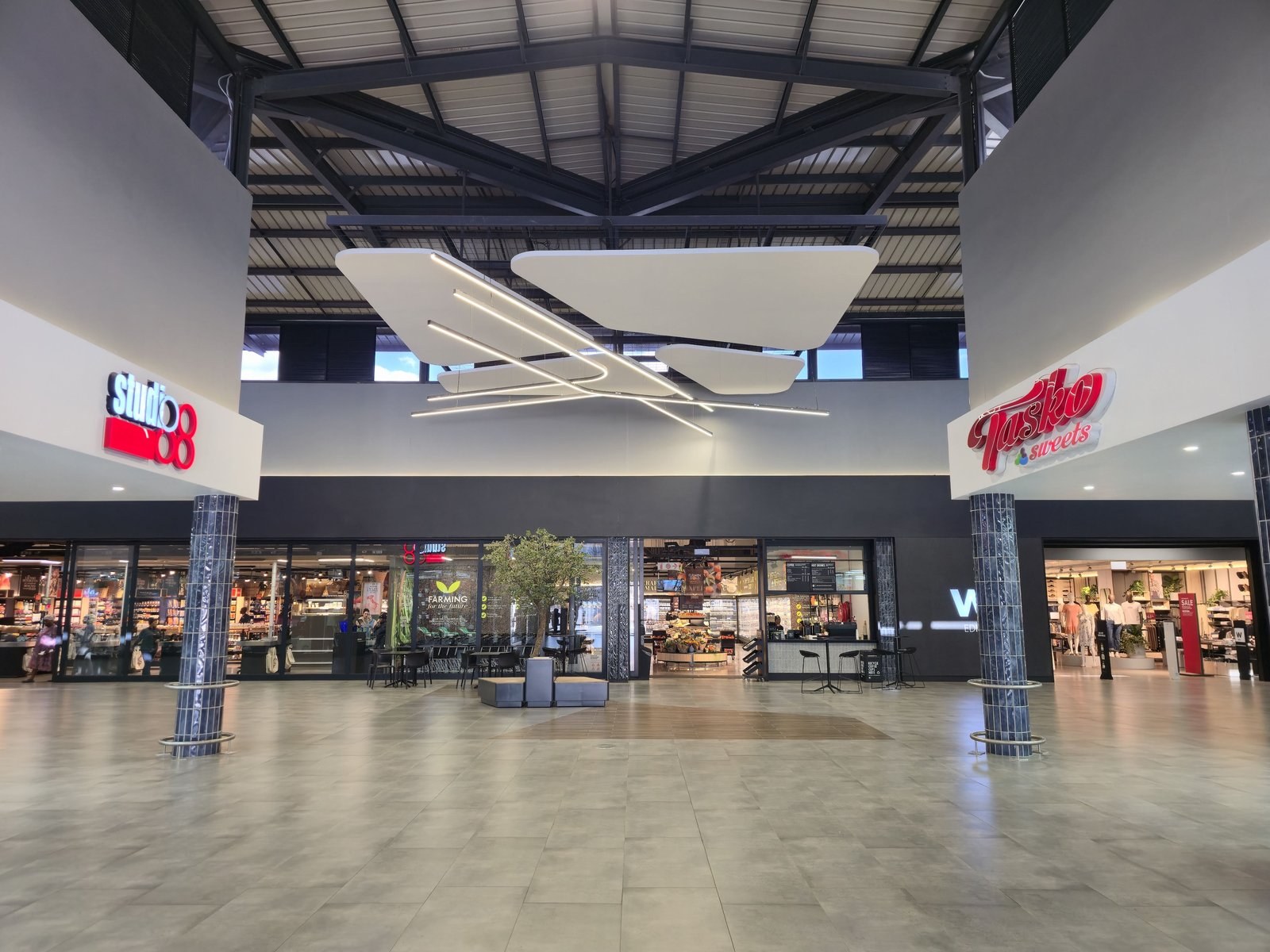Long, slender cut shapes assembled into a hanging sculpture with linear light bars, in the double-volume court beneath an exposed steel roof.