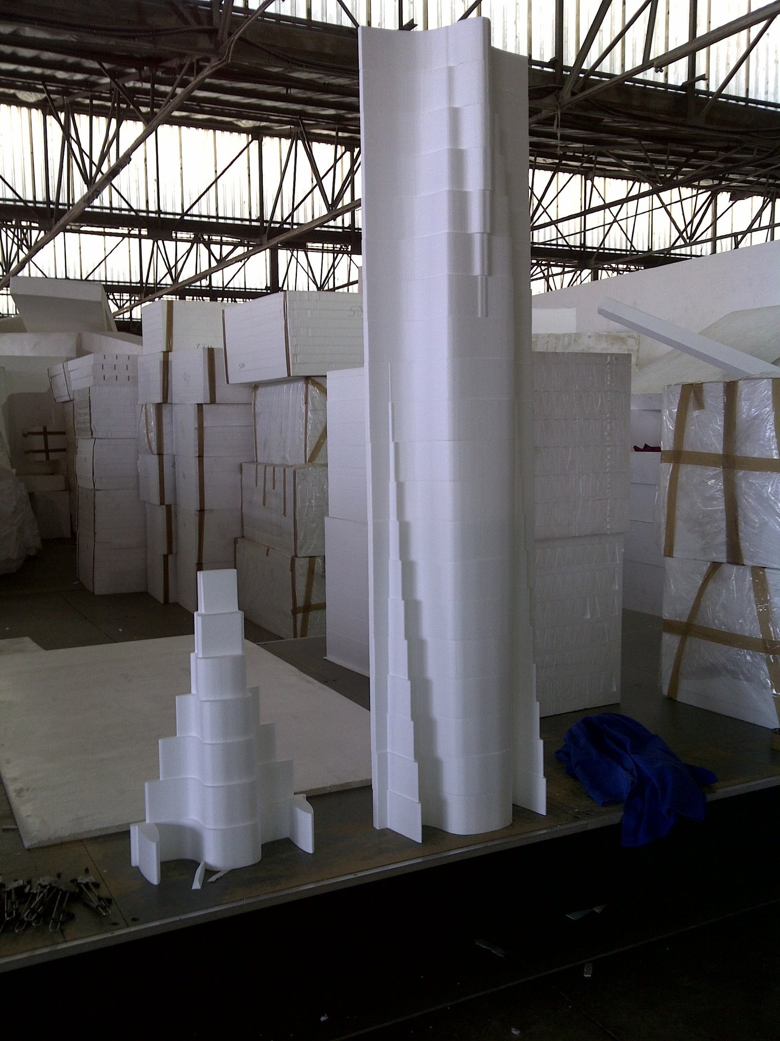 Large sculpted EPS column and a stepped spiral piece on a workbench, with stacked wrapped EPS block inventory in the warehouse behind.