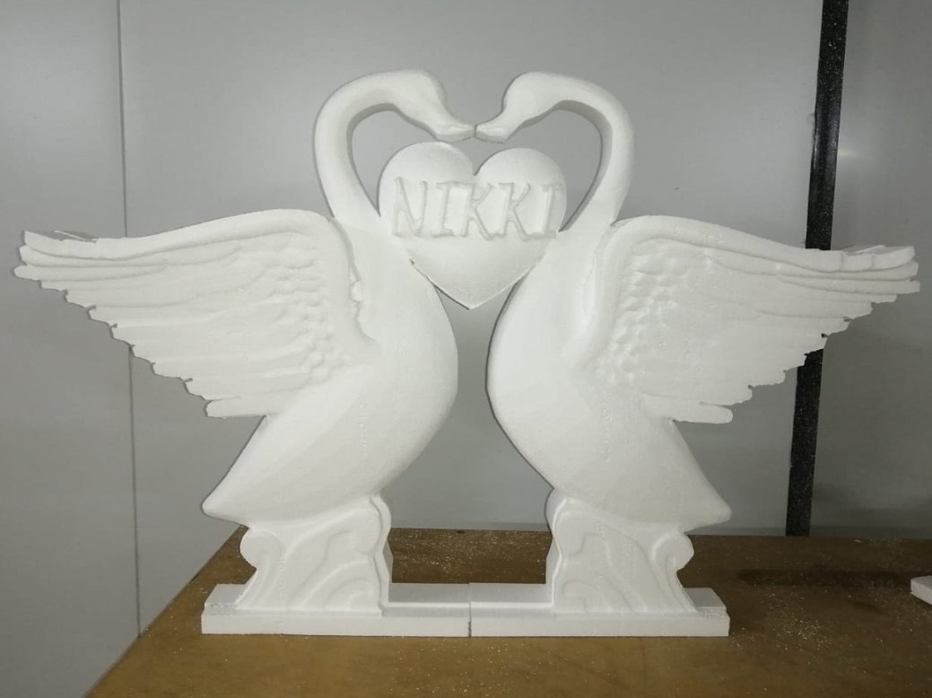 Two EPS swans milled so their necks meet to form a heart, with the name NIKKI carved into the heart panel between them. Stepped feather detail on the wings, scrolled pedestals, and a two-piece base plate; photographed on the workbench with cutting dust still on it.