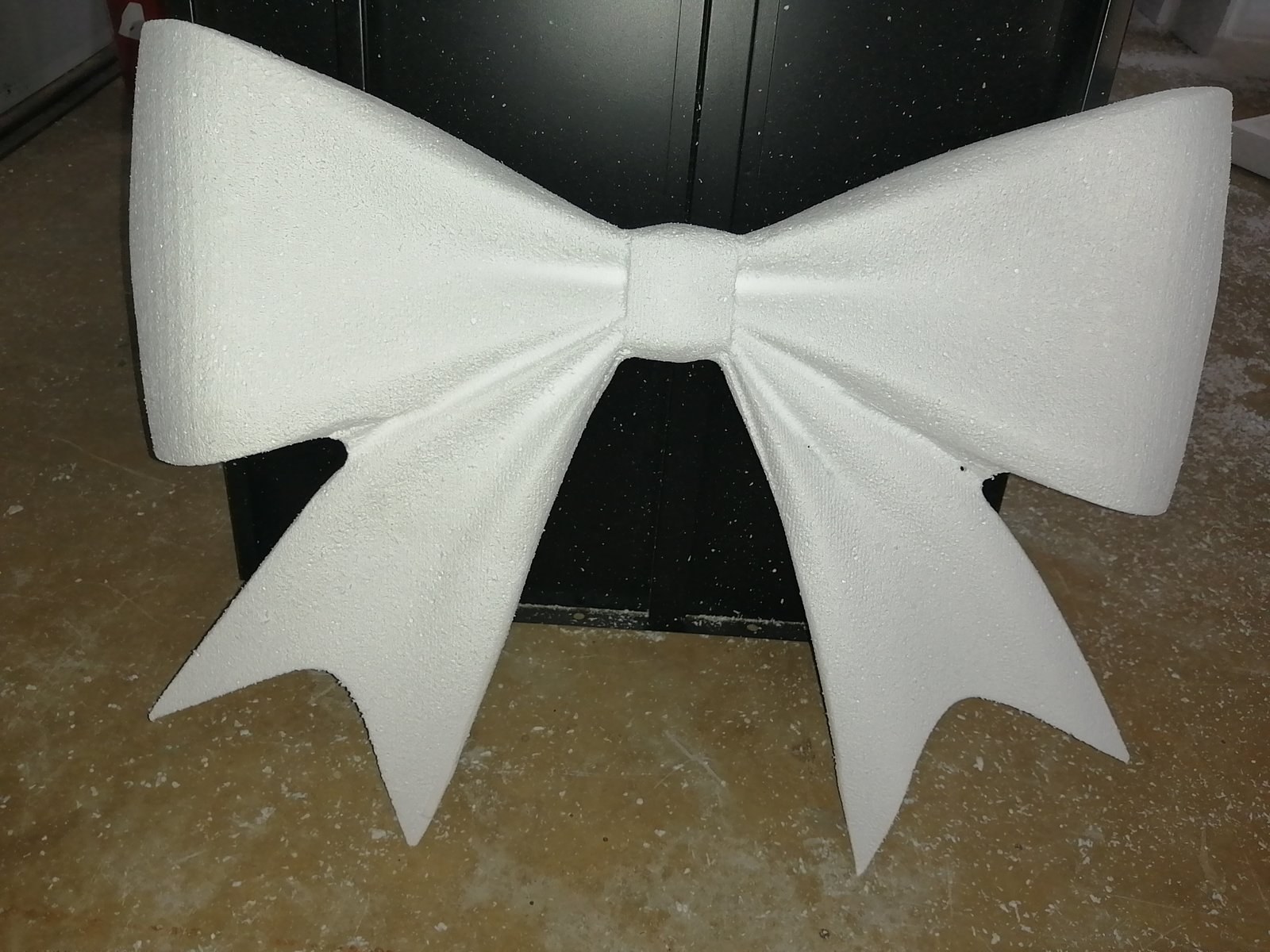 An oversized decorative bow milled from EPS - two loops, a centre knot and swallow-tailed ribbon ends - standing on the workshop floor beside the machine, with cutting debris still around it.