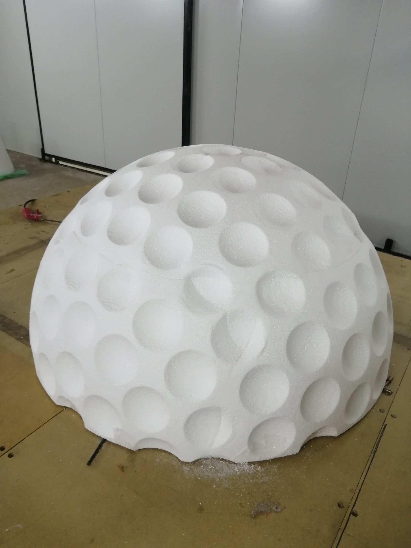 A golf-ball dome milled in EPS, the dimples cut individually into the curved surface. It sits on the machine bed with the join line between the two stacked foam slabs still visible, and fresh milling shavings on the table.