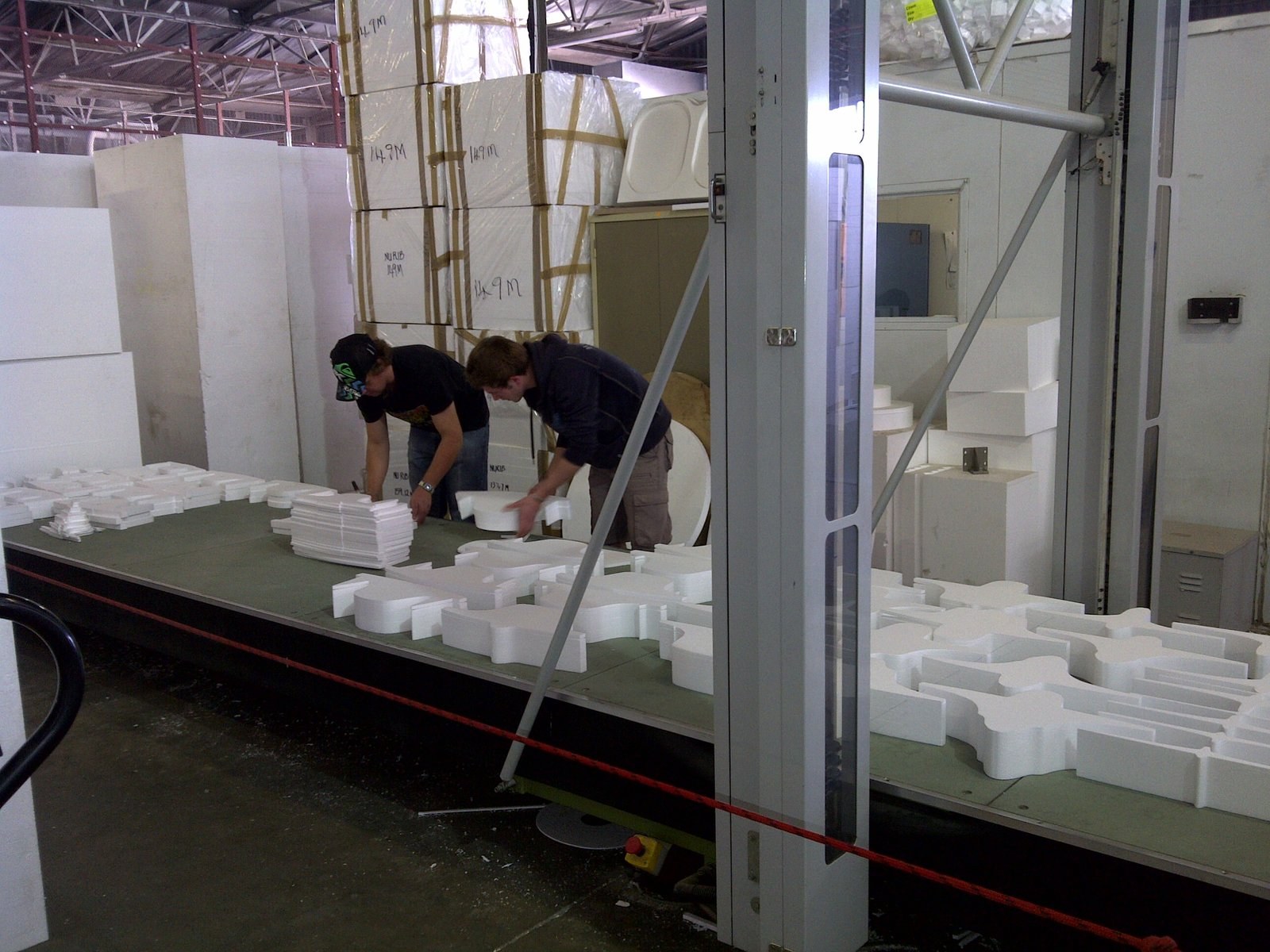 Two workers handling CNC-cut cross-shaped foam pieces on a factory table