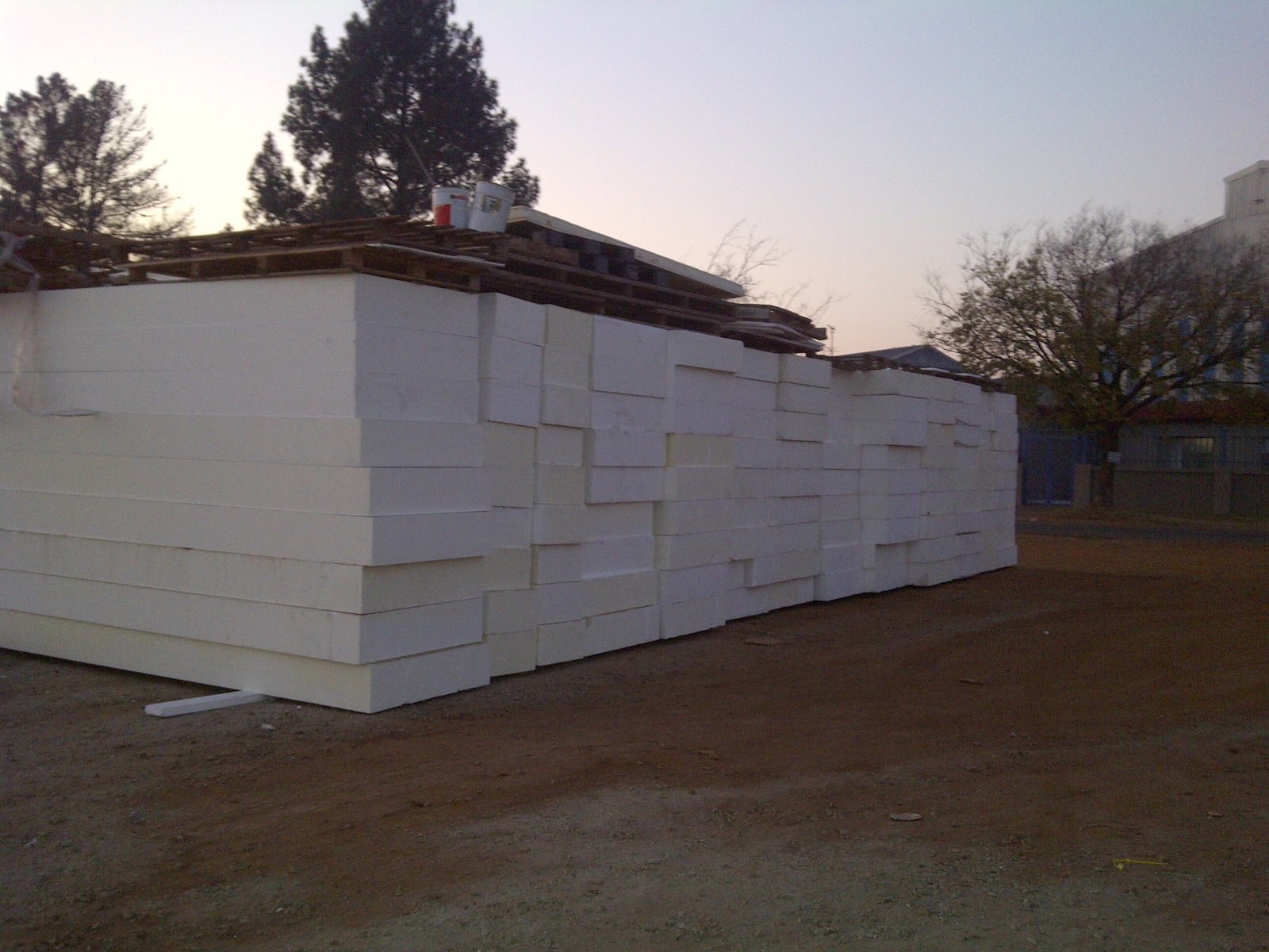 Large EPS slabs stacked several courses high in the yard at dusk, with timber pallets and paint tins left on top. The material in bulk, outdoors, before dispatch.