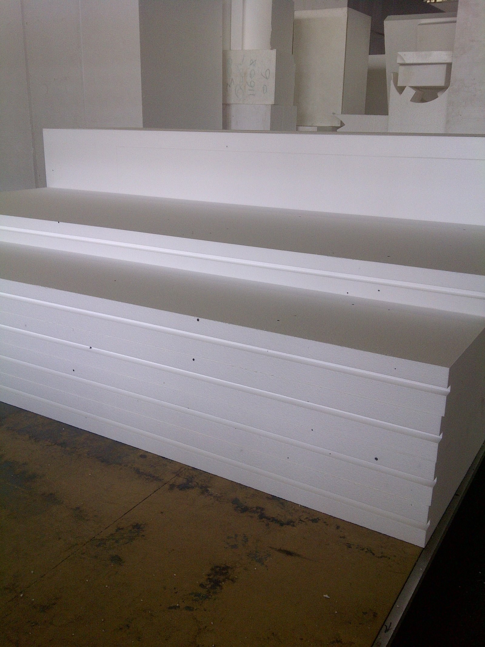 Cut white EPS boards stacked flat on the factory floor, each with a rebated shiplap edge along its long side; hand-marked EPS shapes stand behind them.