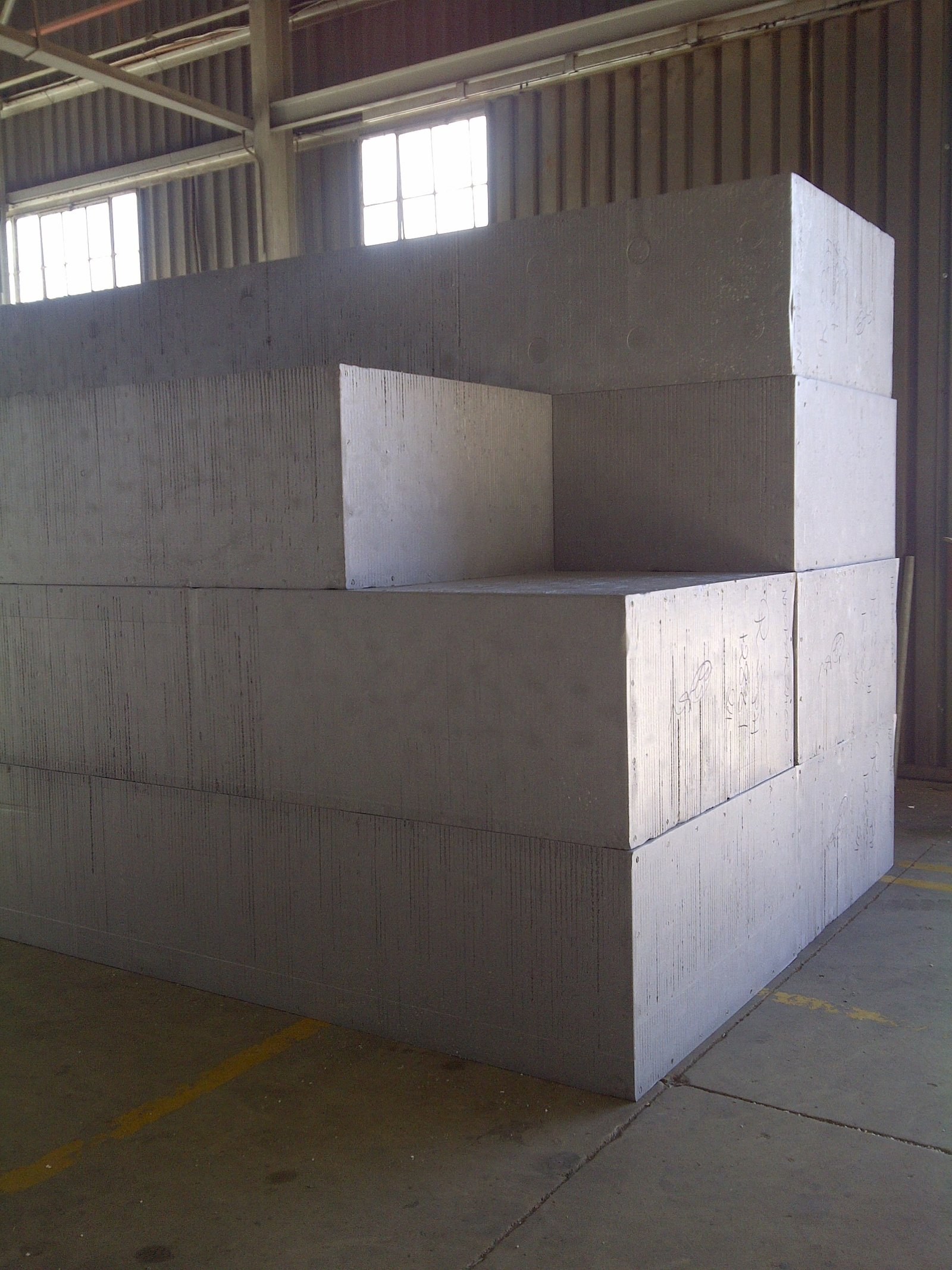 Full-size uncut EPS billets stacked in the factory bay, hand-marked and waiting to be cut down into boards. The vertical ripple on the faces comes off the block moulds.