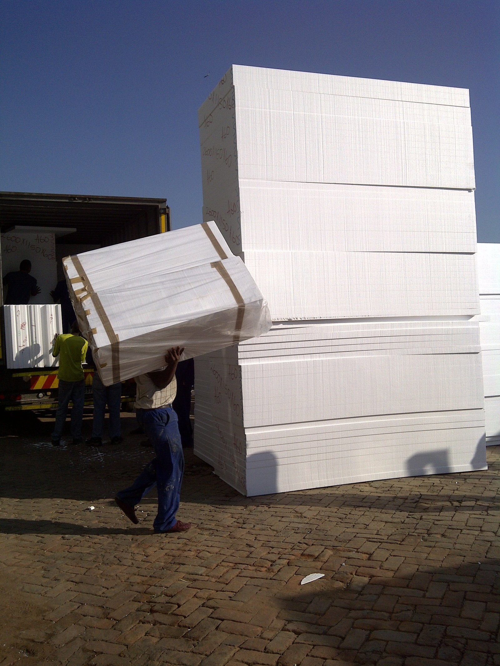 Worker carrying a wrapped EPS board bundle past a stack of blocks in the yard