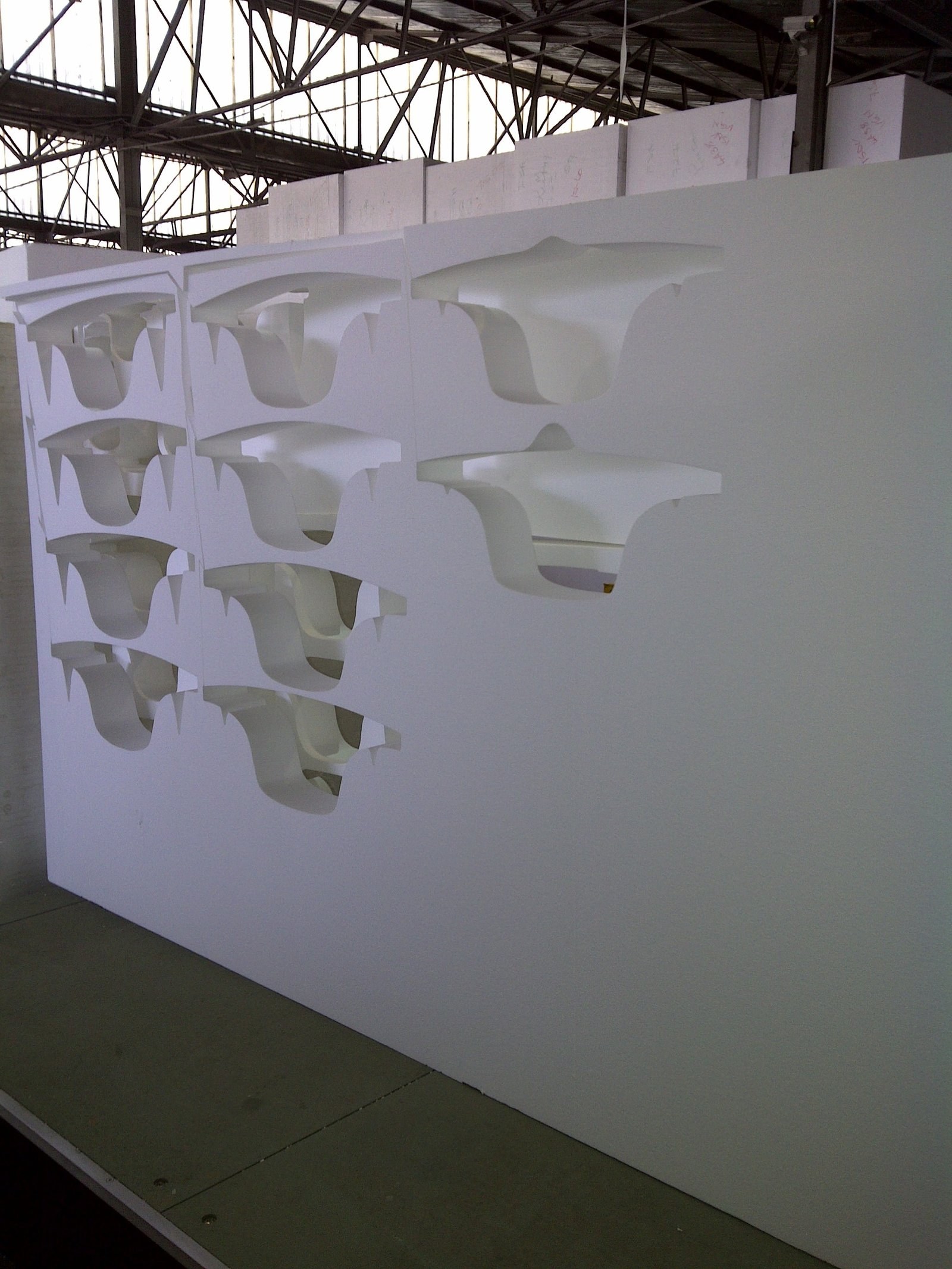 Curved corbel and bracket profiles hot-wire cut through the face of standing EPS blocks, each piece still resting in its own cut-out. Shot on the cutting table under the factory roof trusses; the foam is raw and uncoated at this stage.