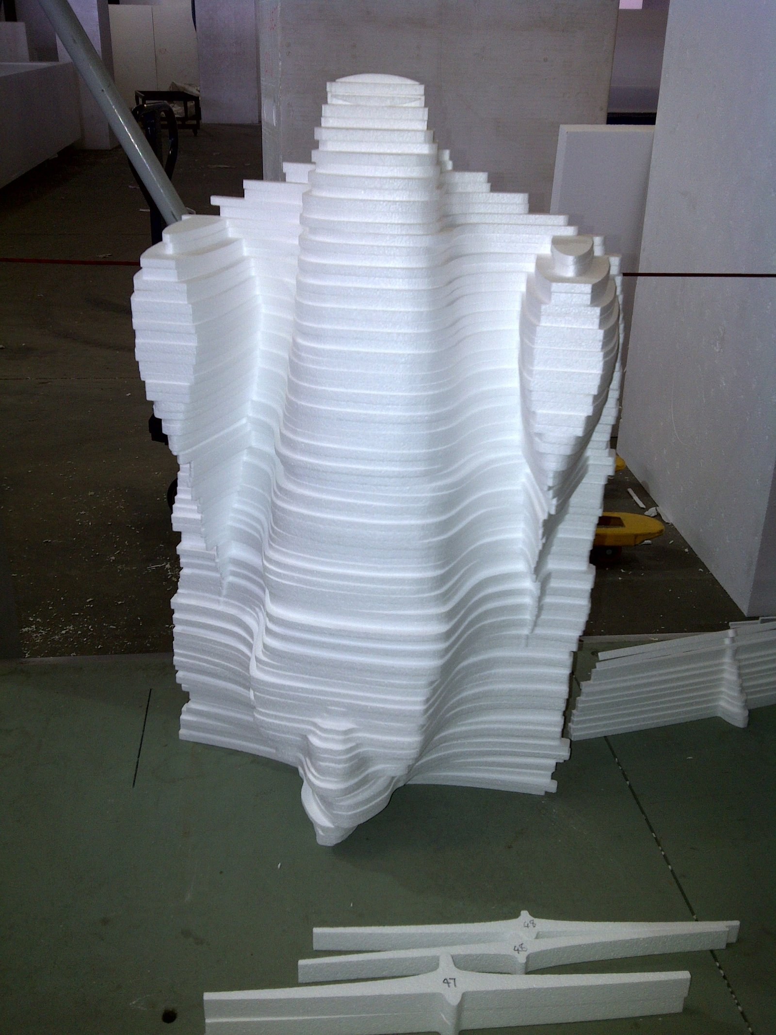 In the factory: a tall decorative element built up from stacked hot-wire-cut EPS layers, the stepped edges forming a fluted shape. Raw uncoated foam, bead texture visible. Numbered cut strips (47, 48) lie on the floor in front.