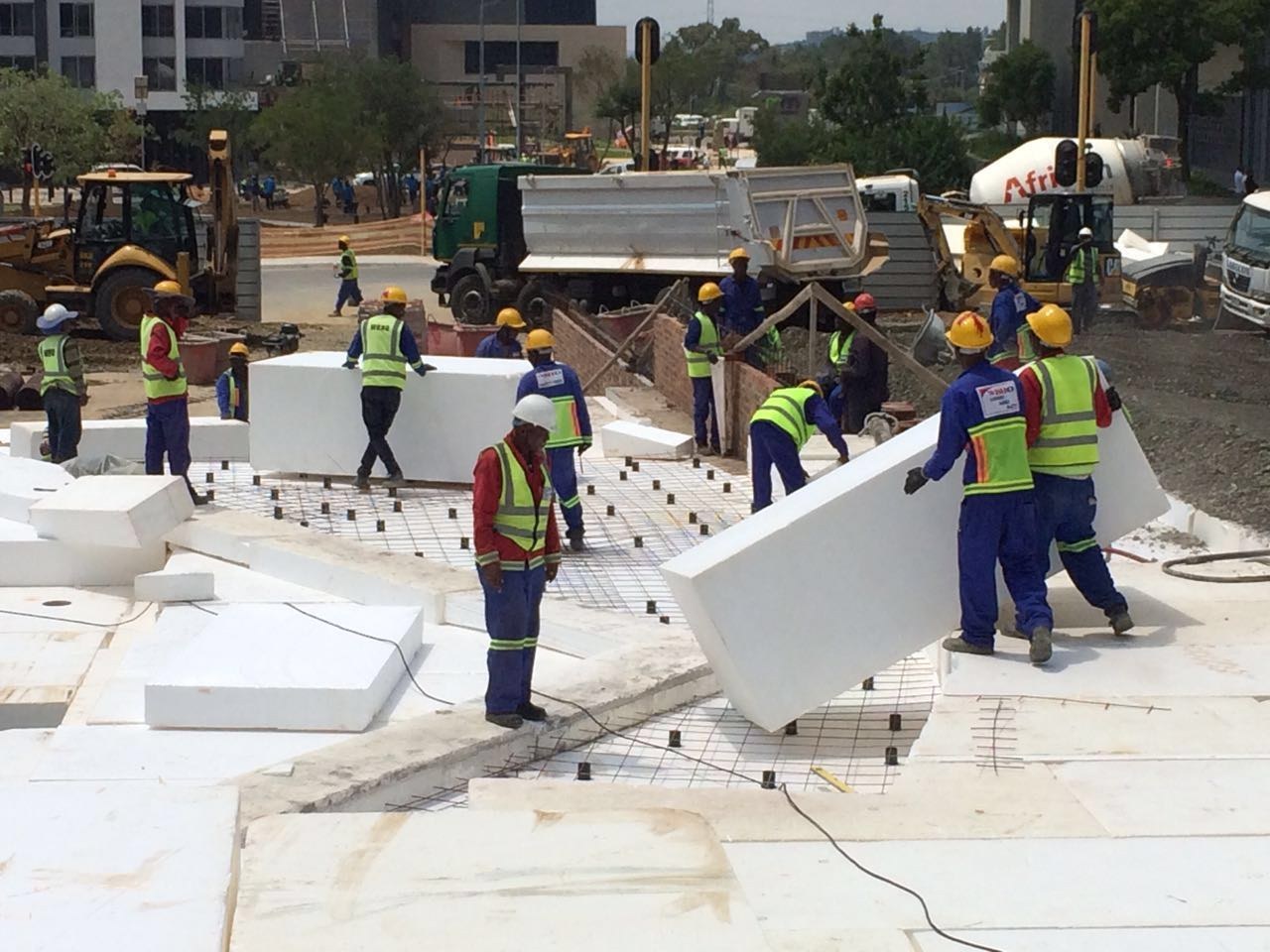 Workers carrying a large EPS block into position on site, with blocks already laid flat and reinforcing mesh on chairs set out over them. Mixer trucks and a TLB working behind.