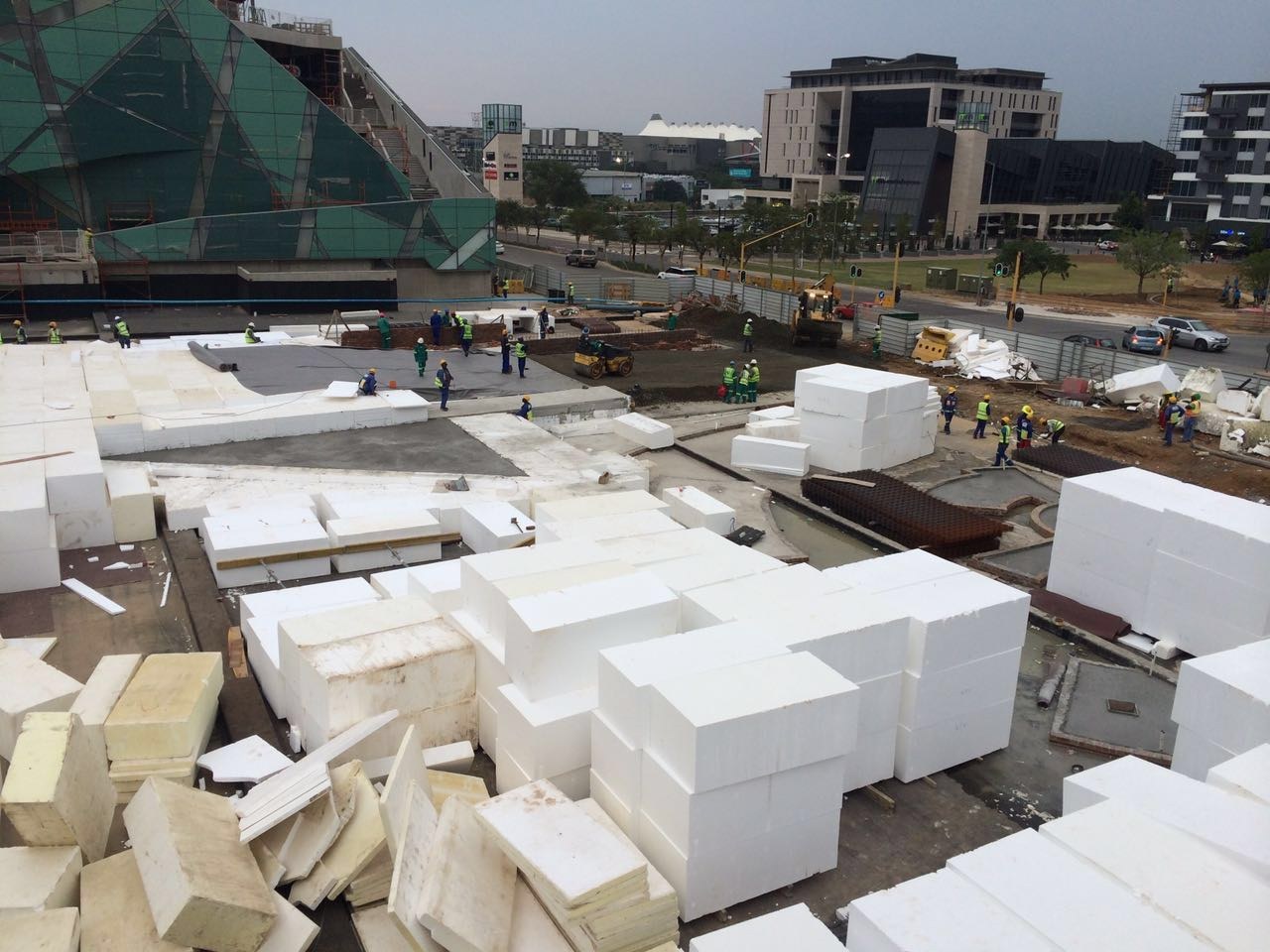 Elevated view of the site with metre-scale EPS blocks stacked in rows across the working area, membrane and reinforcing mesh laid alongside. A roller compactor and crew work between the stacks.