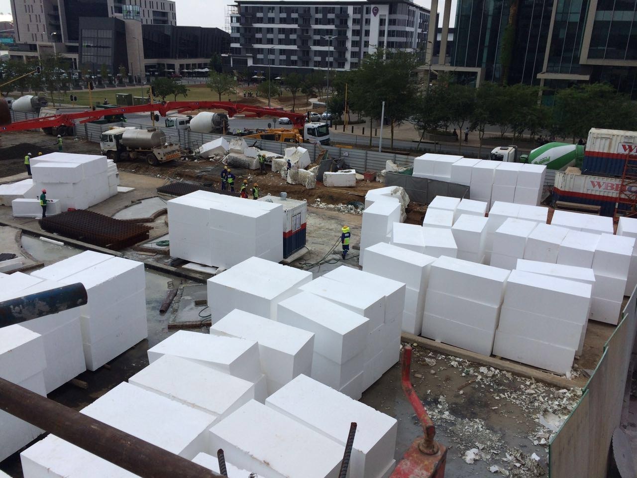 Large EPS blocks stacked several high across the site, staged for placement. A concrete pump boom and mixer trucks stand at the perimeter.
