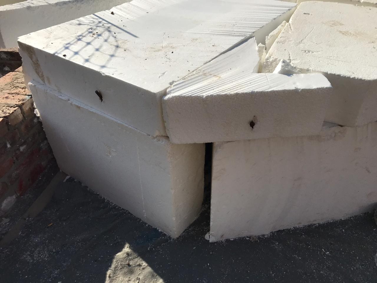EPS blocks butted together over a black waterproofing membrane against a brick wall, showing hot-wire cut faces and steel pins driven in to key adjacent blocks together.
