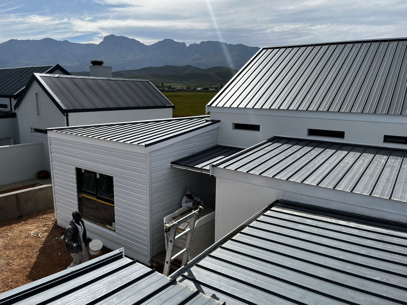 Standing-seam metal roof sheeting laid across several roof planes, with a lower extension clad in white horizontal-lap profiled sheeting. Photographed from the roof during installation, mountains in the background.