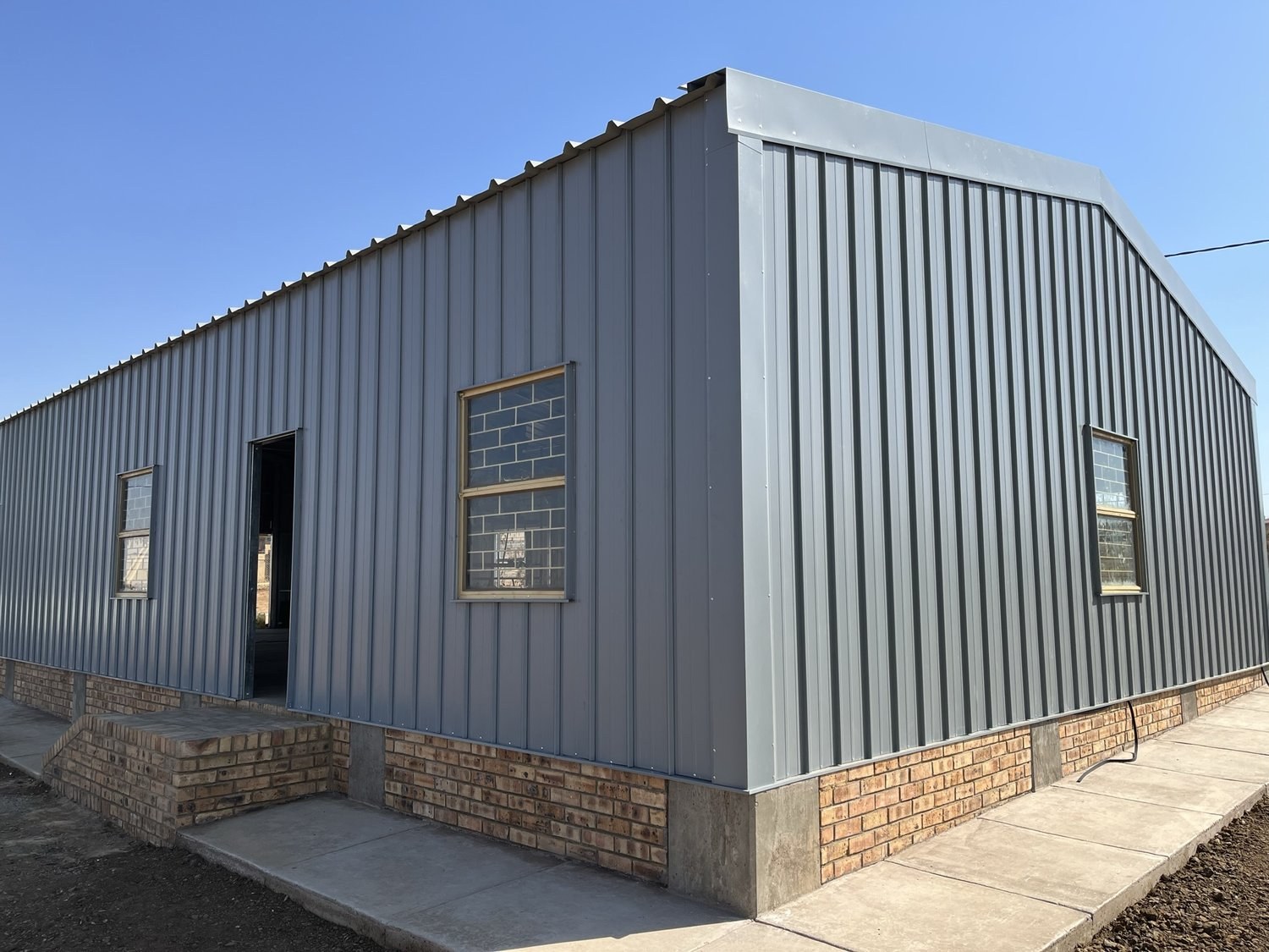 Building fully clad in grey vertical-rib profiled steel sheeting, with folded barge caps at the roof edge and a face-brick plinth at the base.