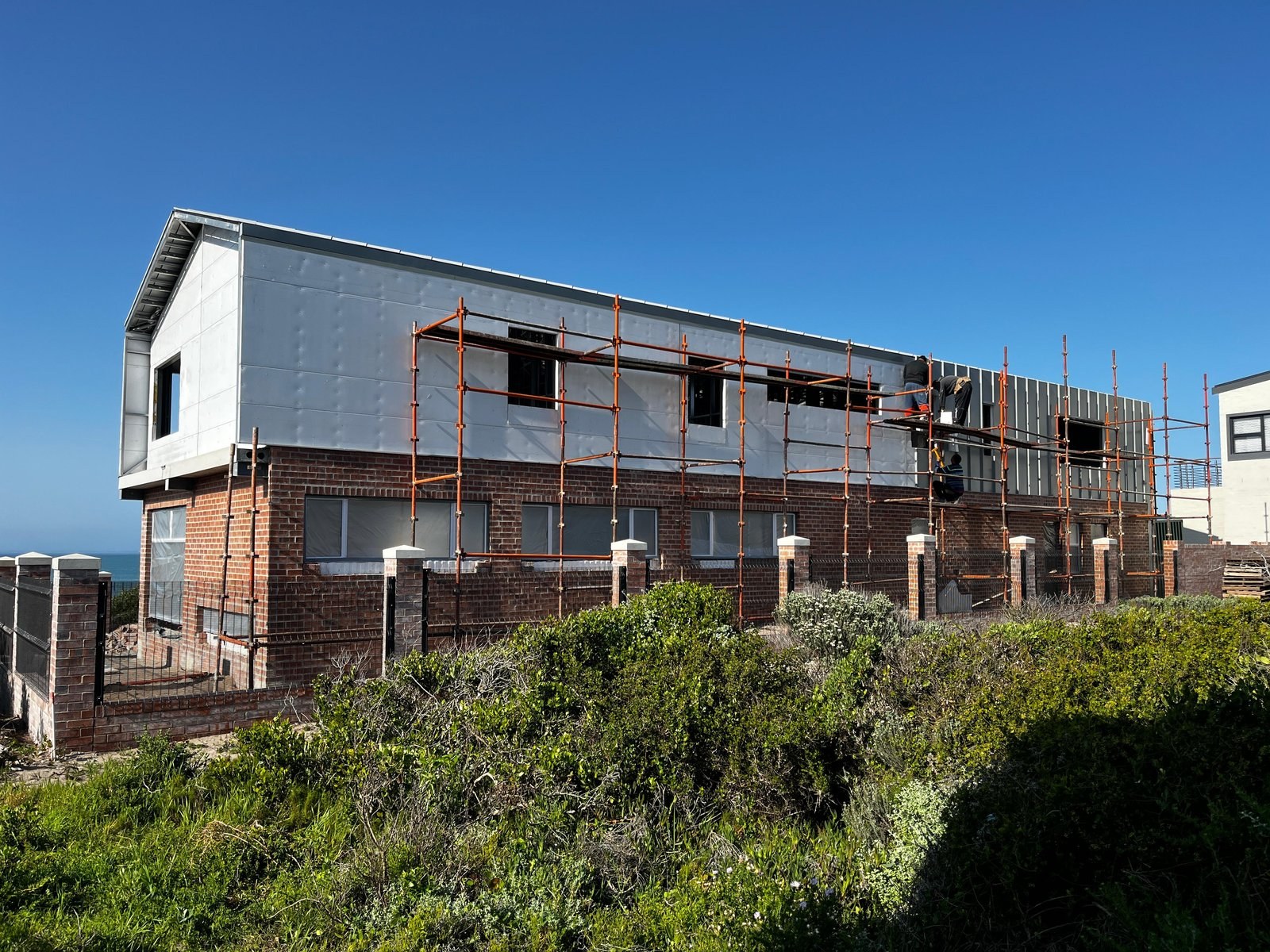 Steel wall cladding partway through installation over a brick upper storey of a coastal house, scaffolding in place