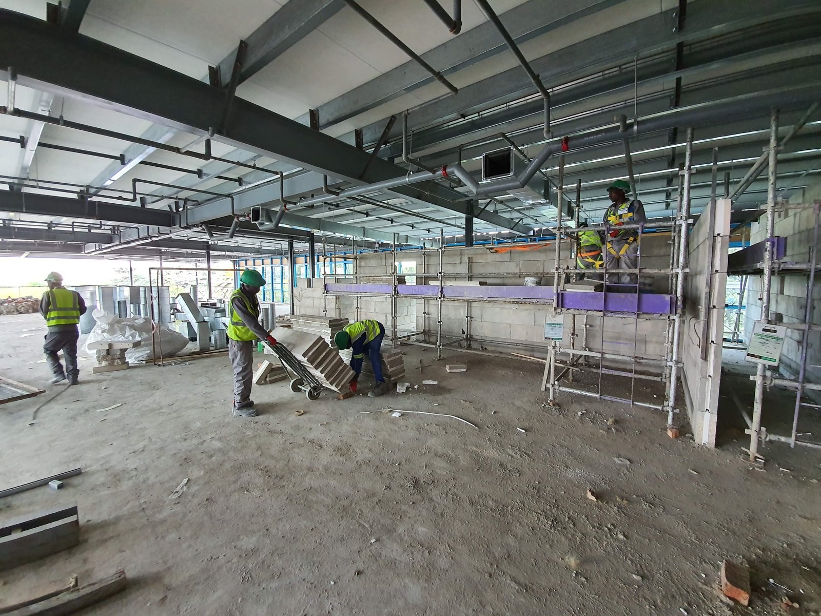 Workers moving panels on a trolley and laying blocks from scaffold inside a steel-frame building.