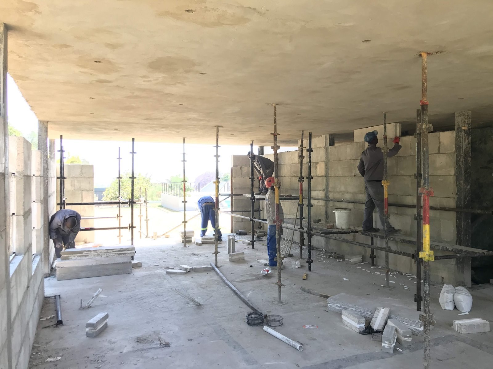 Workers building block walls on adjustable steel props inside an open house shell.